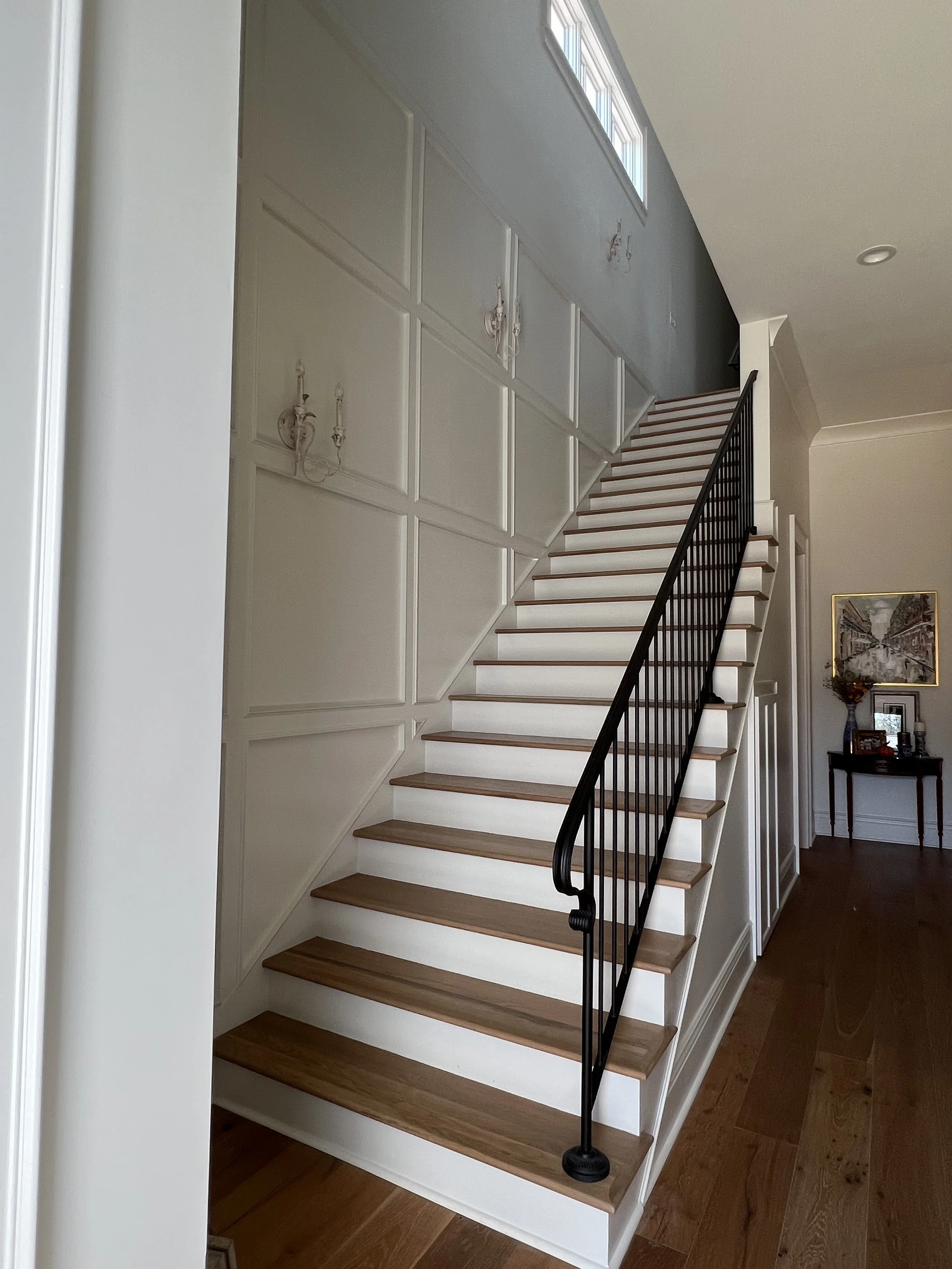 Indoor wooden staircase with white risers and black metal railing, adjacent to a white paneled wall with wall sconces, in a home interior.