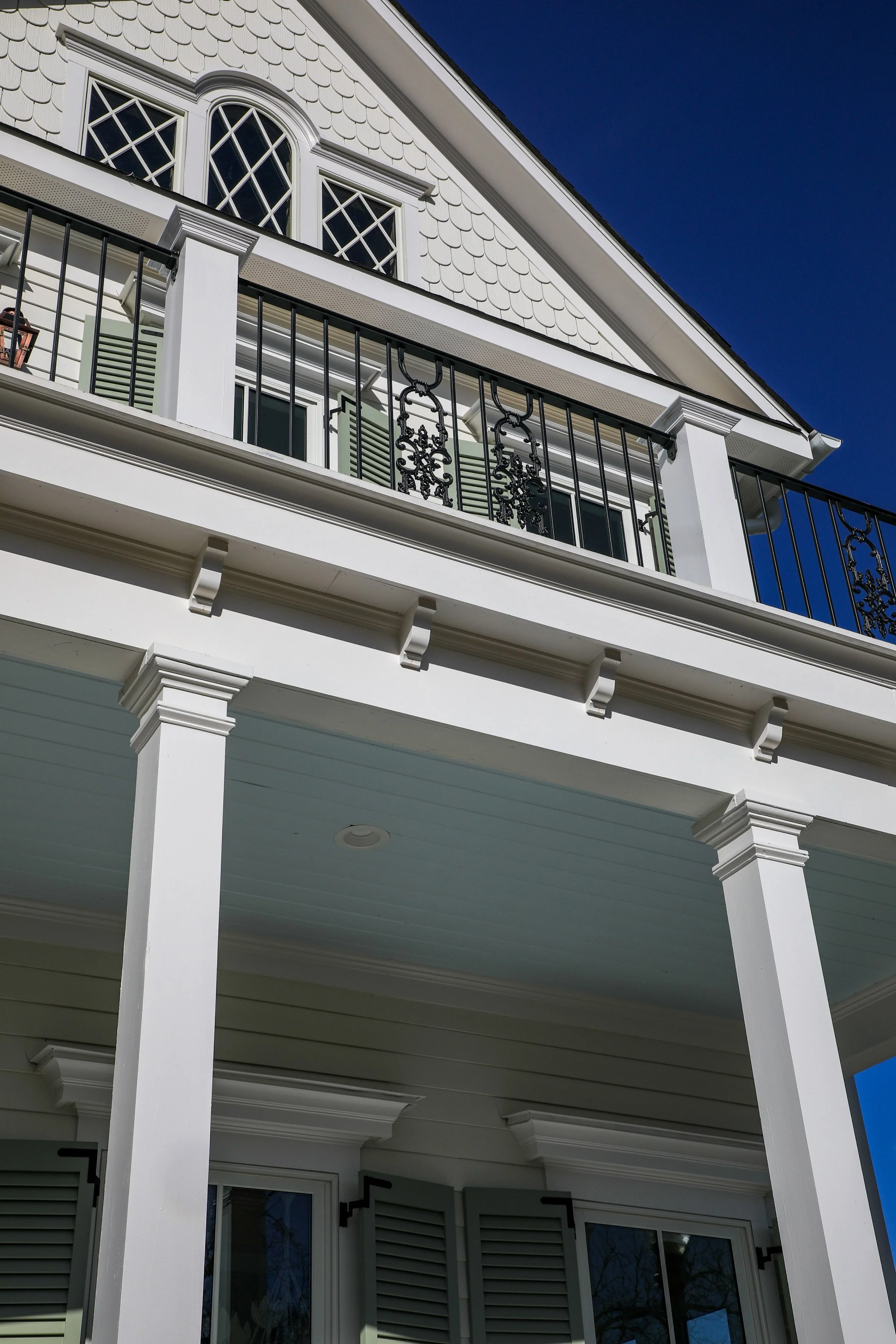 Looking up at the exterior of a white three-story house with a balcony, decorative trim, and a gabled roof against a clear blue sky.