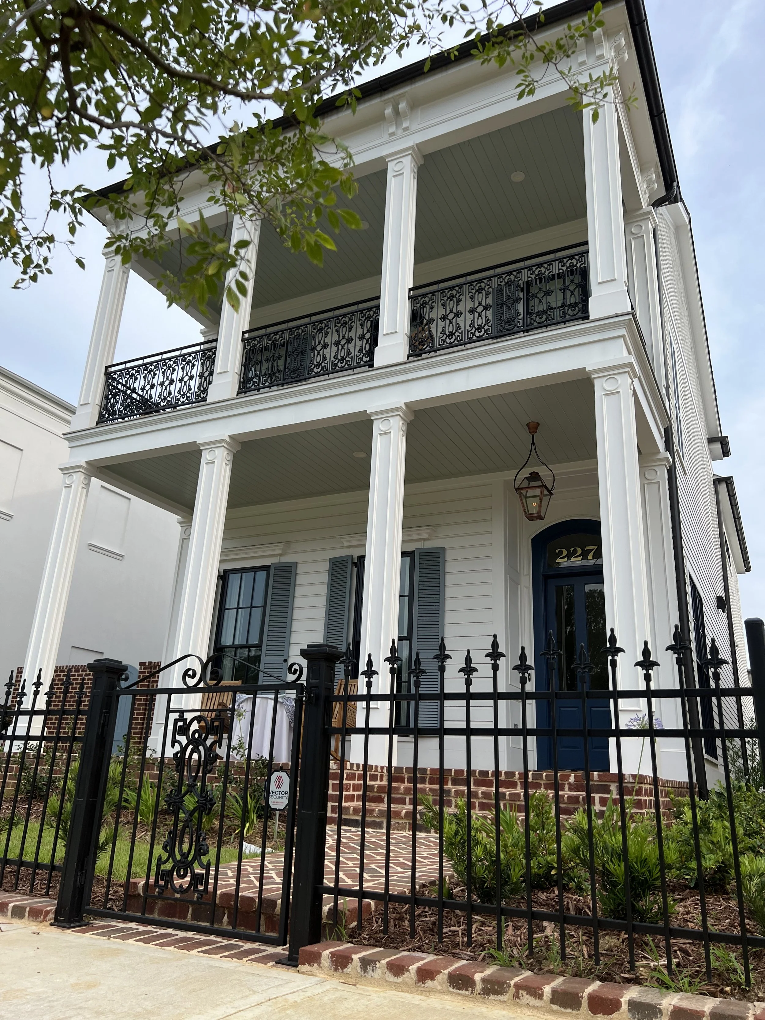 Two-story white house with blue door, black wrought-iron balcony, and columns, surrounded by a black metal fence, with brick steps and plantings in the front yard.