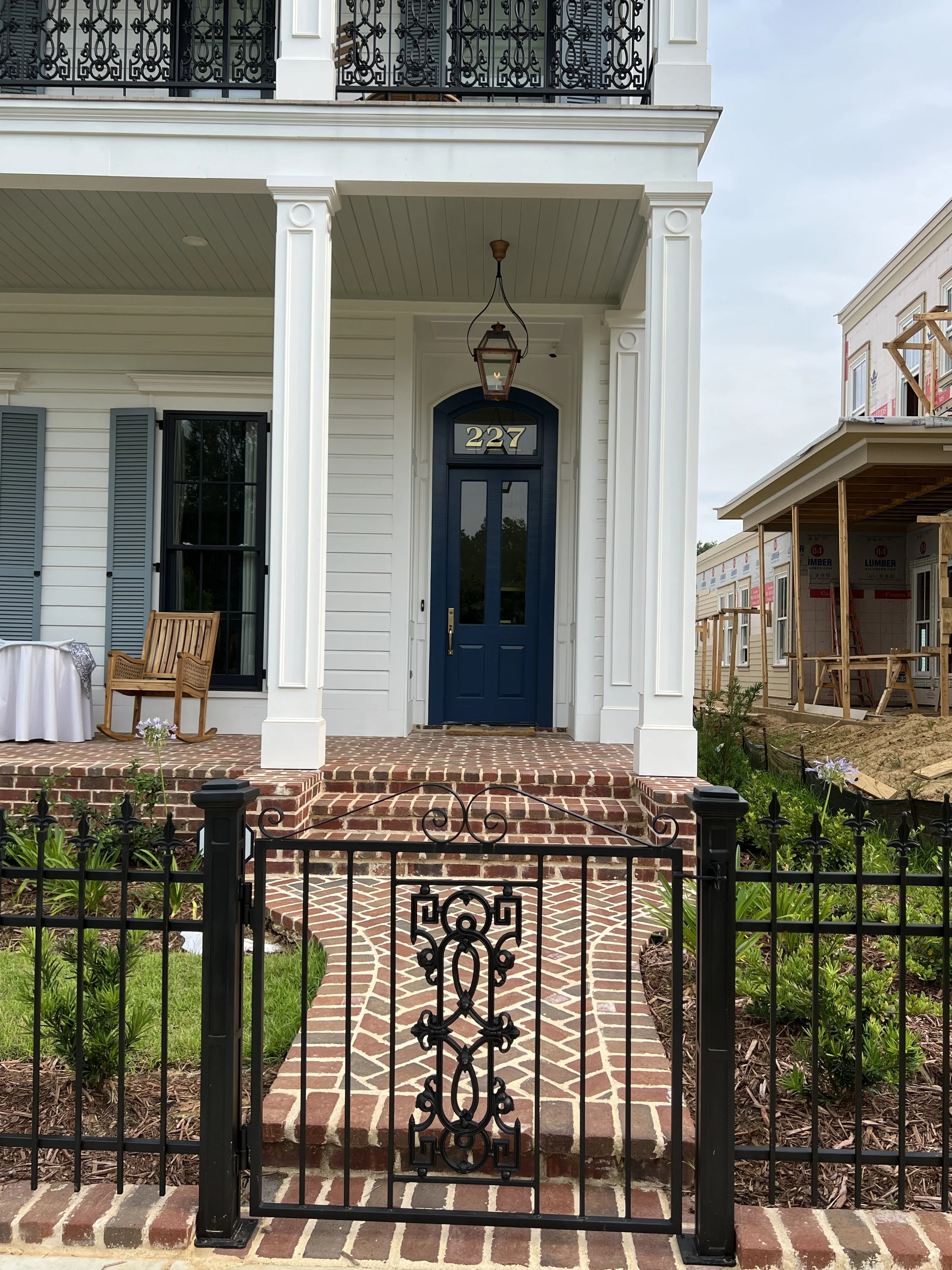 Front entrance of a house with a brick walkway, black metal gate, white siding, blue door, and hanging lantern light, with a porch adorned with wooden benches and plants.