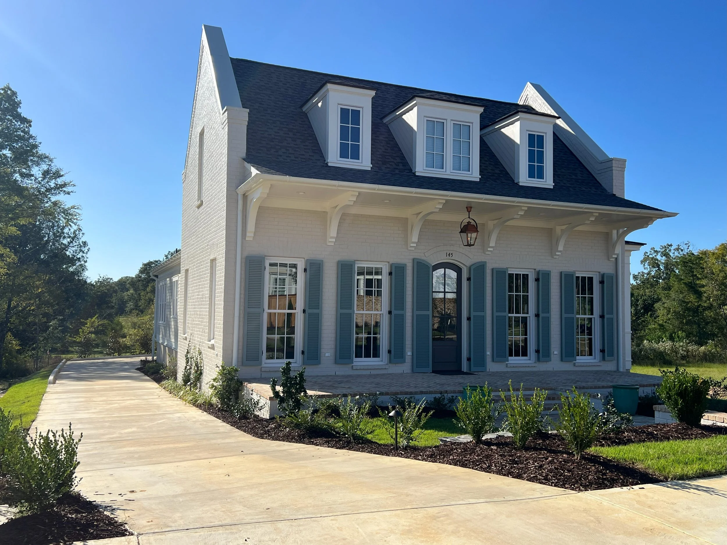 A white two-story house with blue shutters, a black roof with three dormer windows, and a front porch with decorative braces and a hanging lantern. There is a walkway leading to the house and a landscaped yard with small plants and bushes.