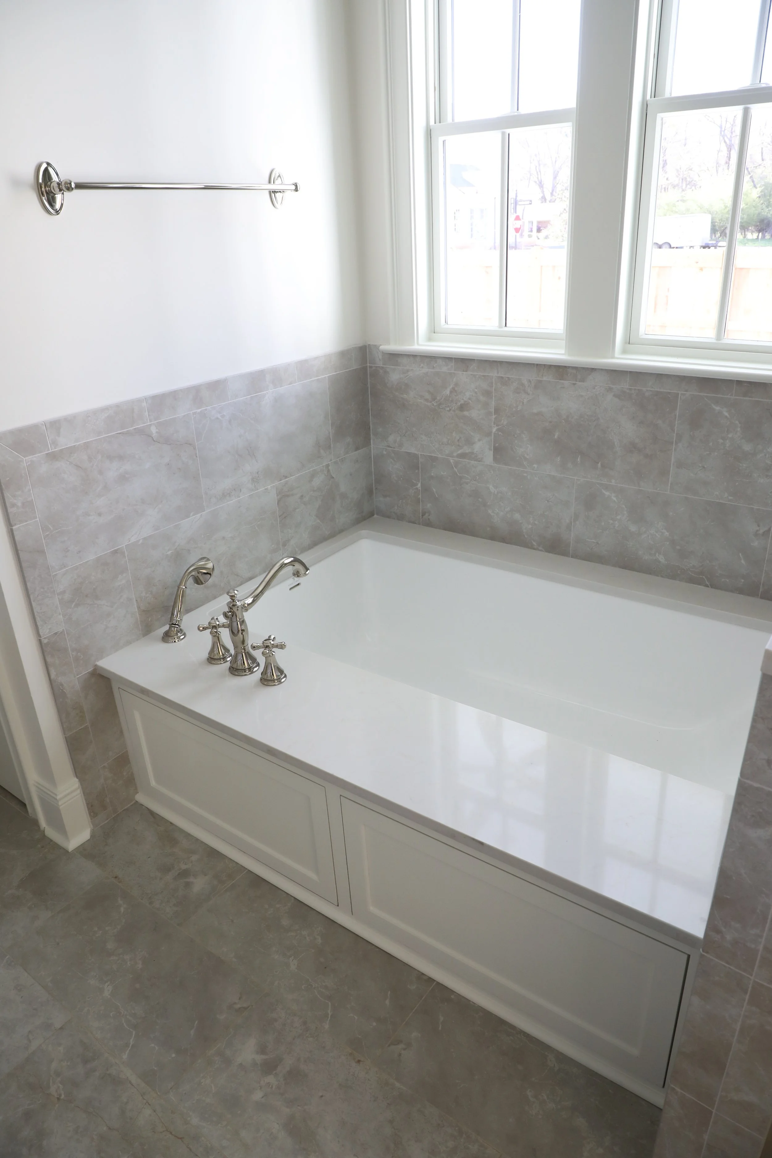 White bathtub with silver fixtures in a bathroom, surrounded by gray tiles, next to a window with white framing.
