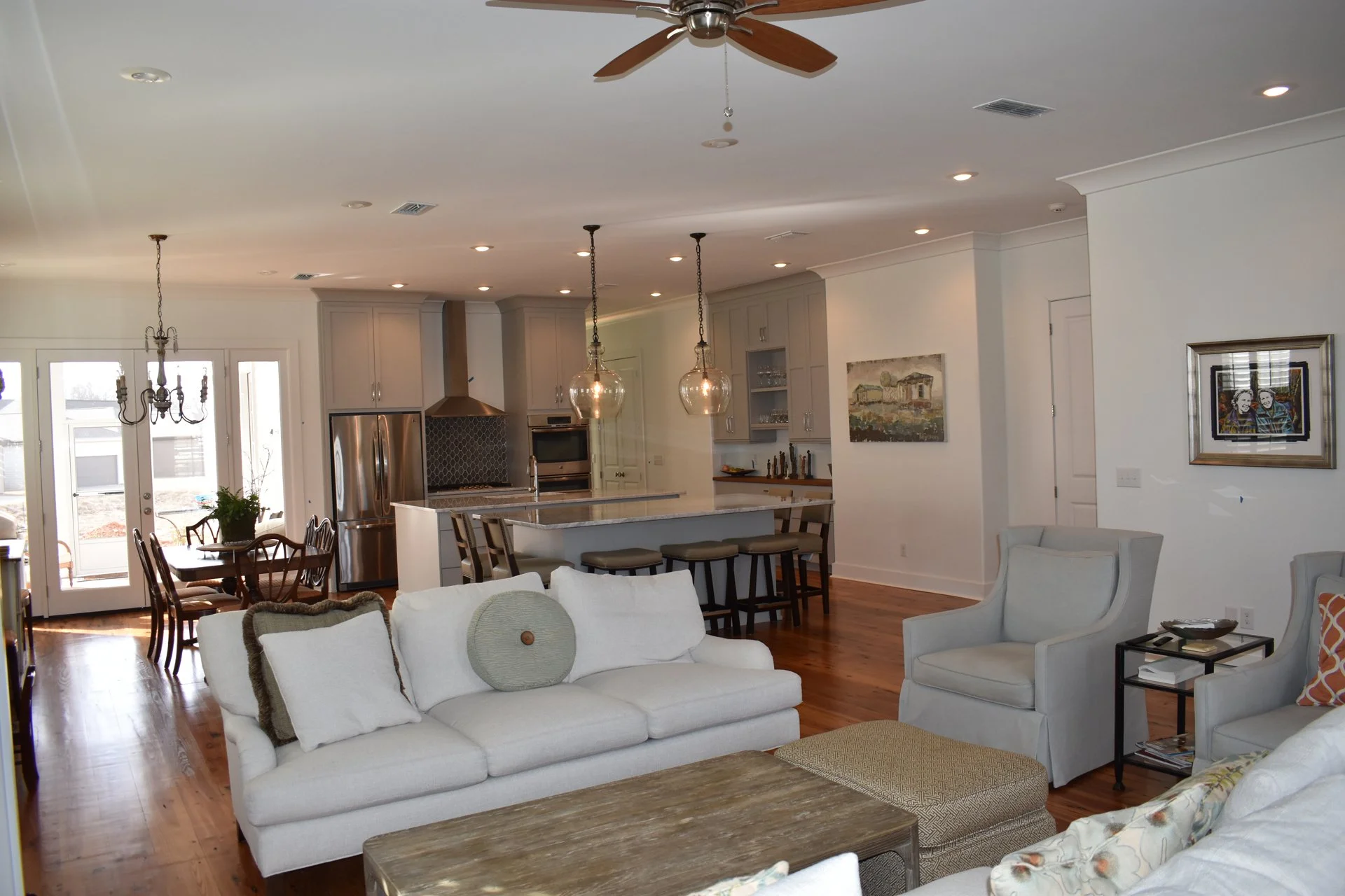 Living room and kitchen area with white sofa, armchair, dining table, kitchen island, pendant lights, and stainless steel appliances.