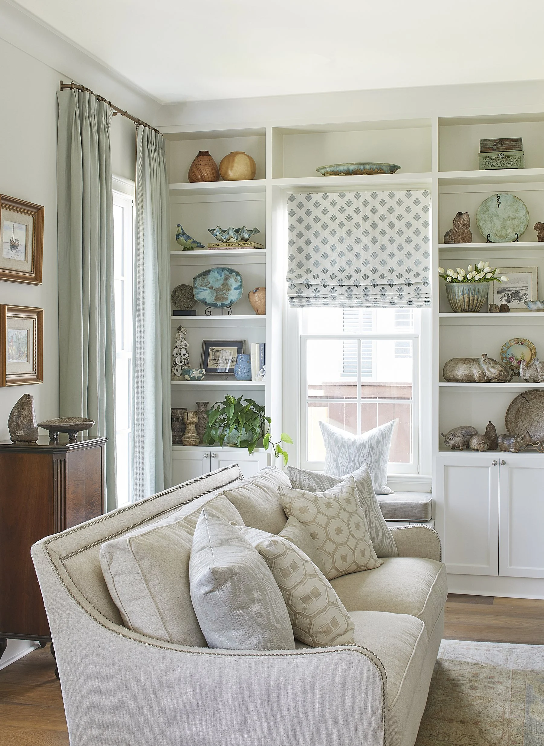 Living room with a beige sofa, decorative pillows, built-in white shelves filled with ceramics, vases, and artwork, a window with a patterned roller blind, and curtains, in a bright and airy space.