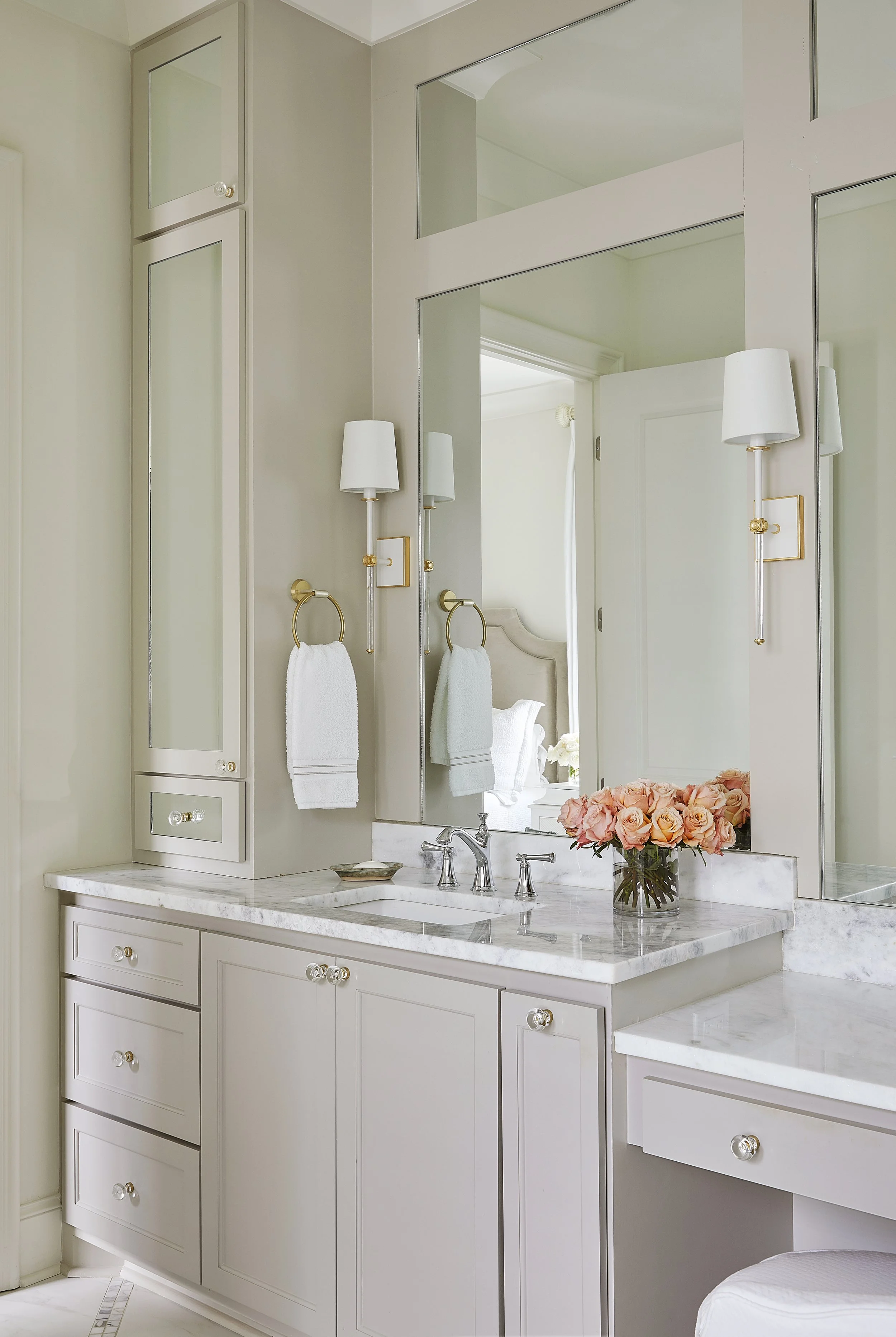 A bathroom vanity with a white marble countertop, matching cabinets, a rose bouquet, and a large mirror. Two wall sconces with white shades flank the mirror. Towel rings with white towels are mounted on the wall.