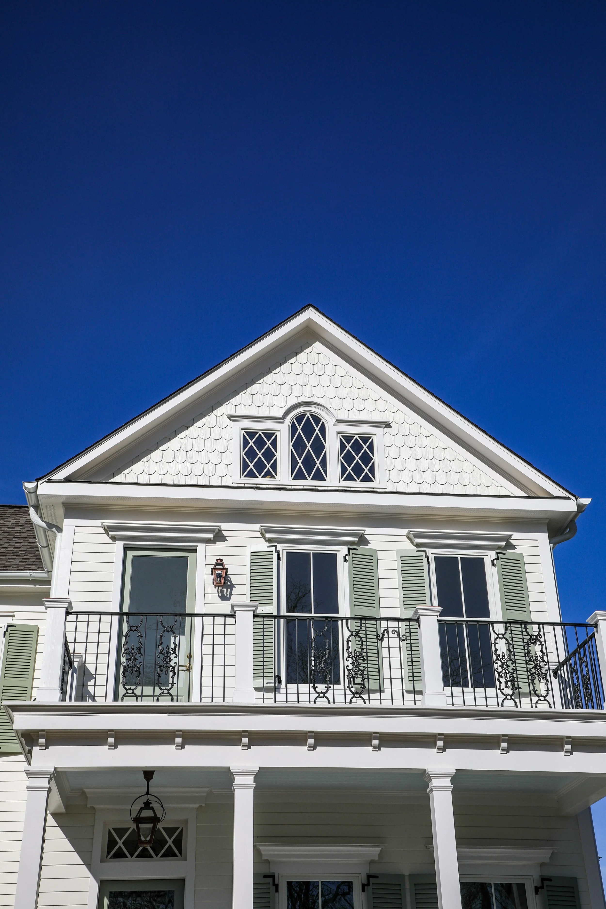 Front view of a white house with a balcony, green shutters, and decorative window panes against a clear blue sky.