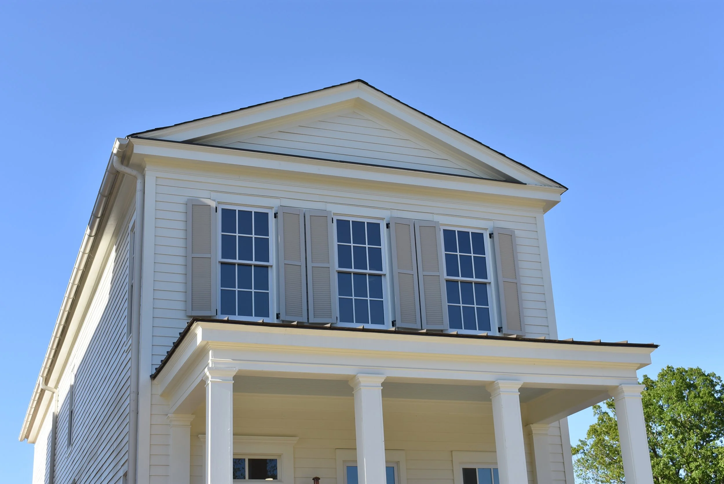 Front view of a white, two-story house with three windows and shutters on the second floor, a porch with columns, and a black roof, against a clear blue sky.