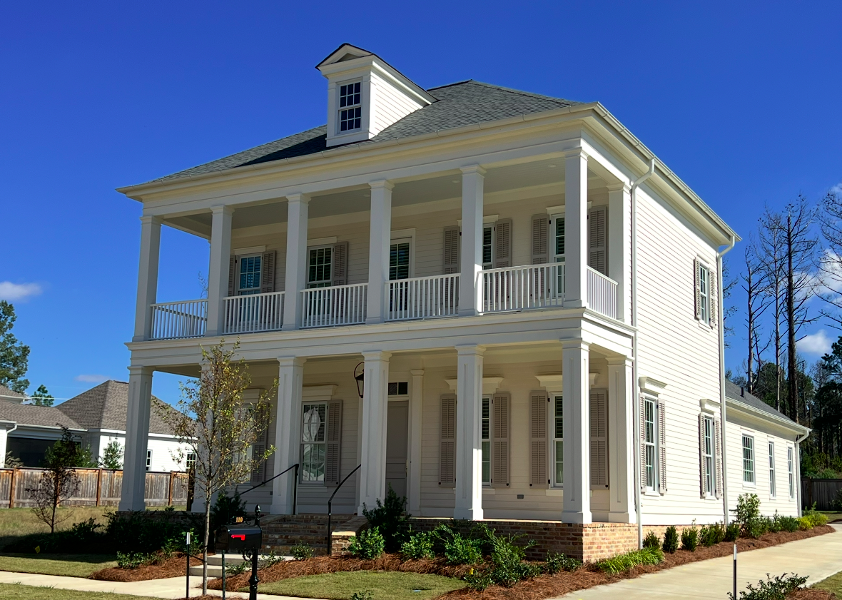 Two-story white house with a porch and columns, black shutters, surrounded by a landscaped yard and a wooden fence, under a blue sky.