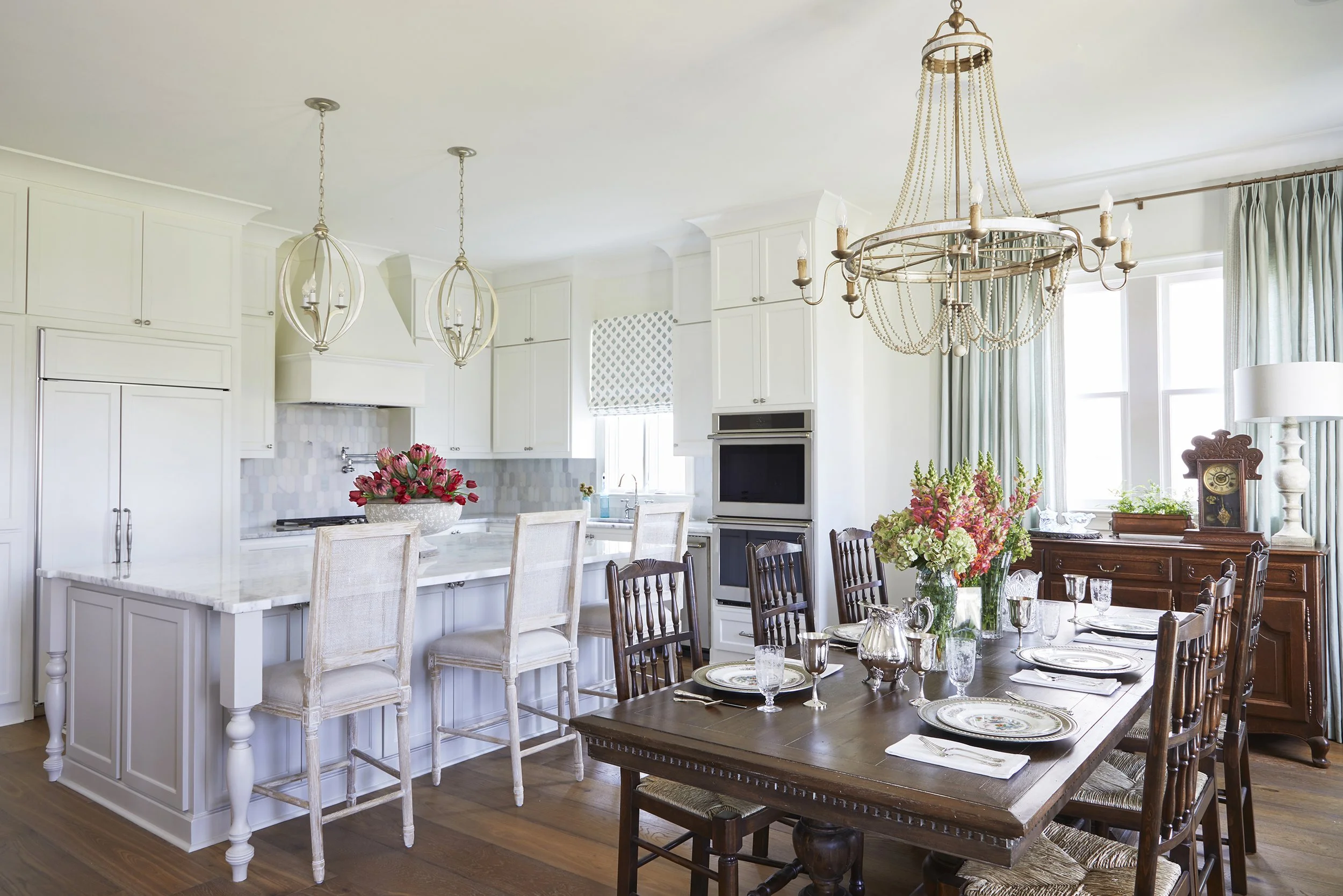 Dining room with dark wood table and chairs set for a meal, floral centerpiece, and chandeliers hanging from ceiling; kitchen area with white cabinets, marble island, and bar stools.