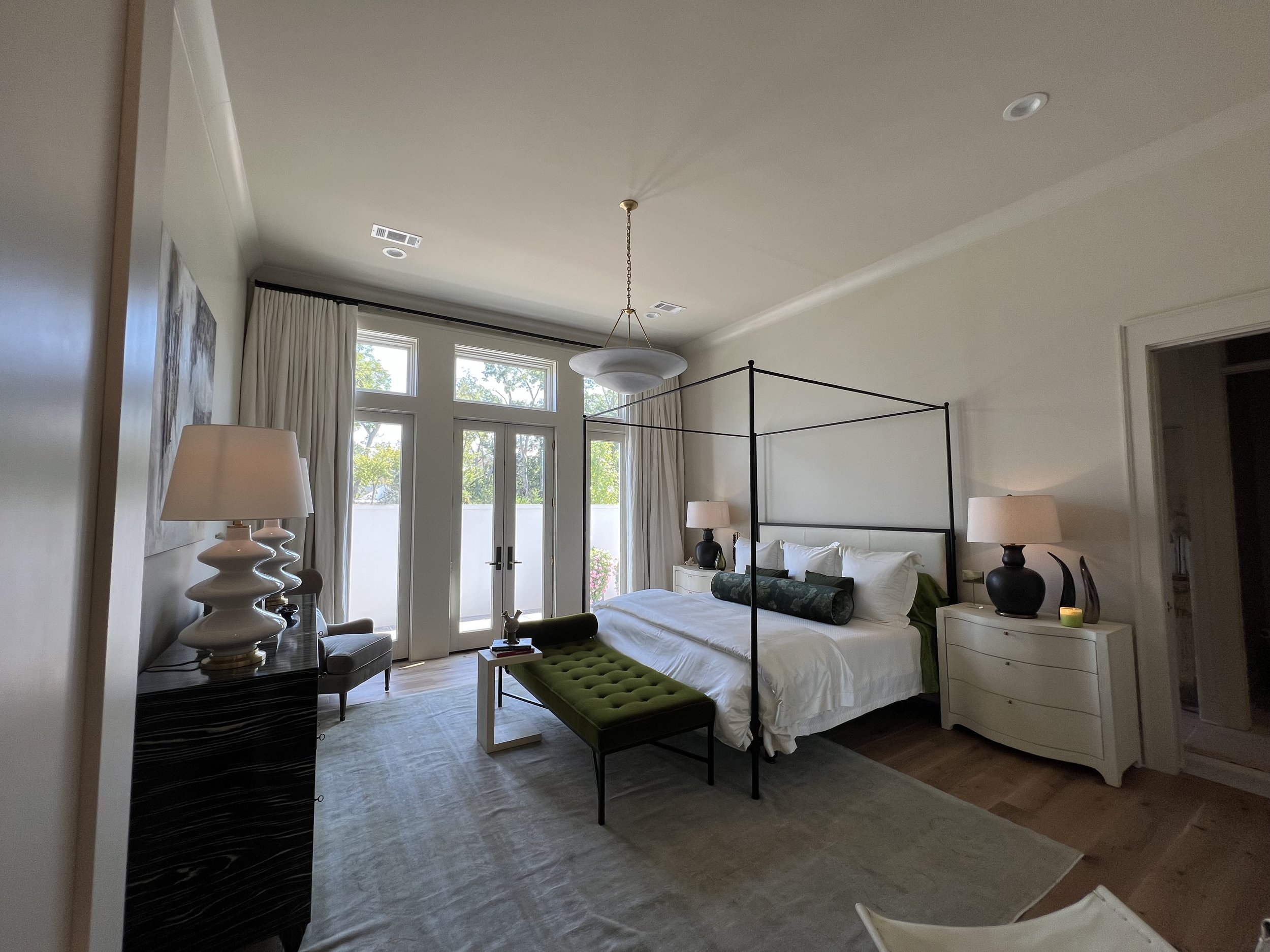 A spacious bedroom with a four-poster bed, white bedding, and decorative pillows. There are two white nightstands with black lamps, a green bench at the foot of the bed, and large windows with white curtains that let in natural light. The room has be