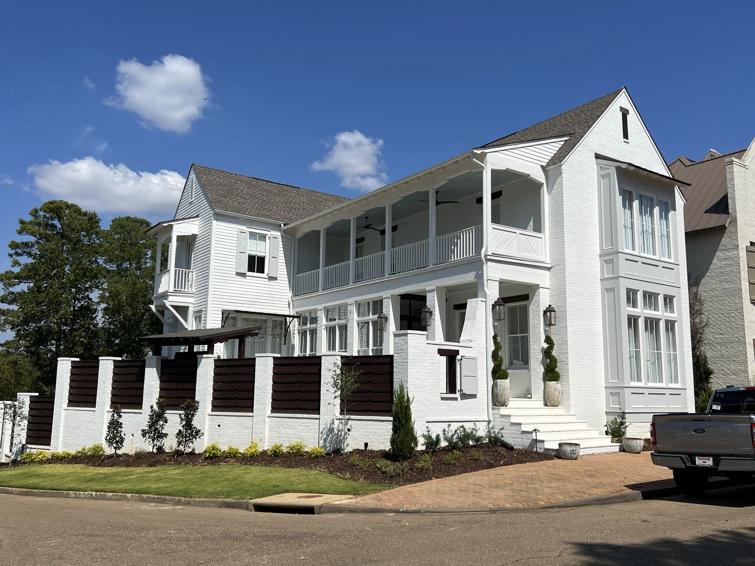 White multi-story house with a front porch, large windows, potted plants, and a brick driveway under a clear blue sky.