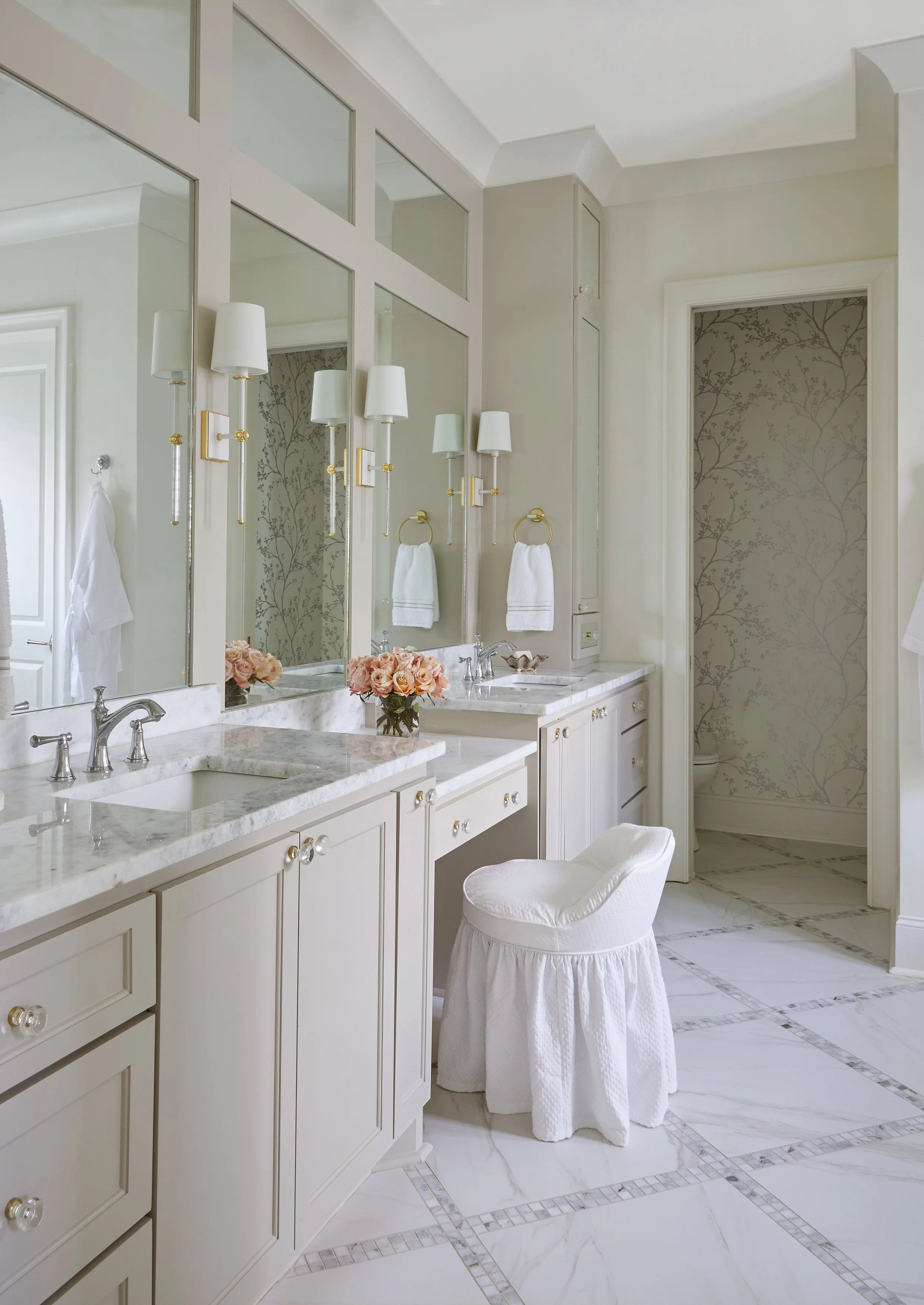 Bright, elegant bathroom with double vanity, marble countertops, large mirrors, gold fixtures, pink roses in a vase, and a white upholstered chair.