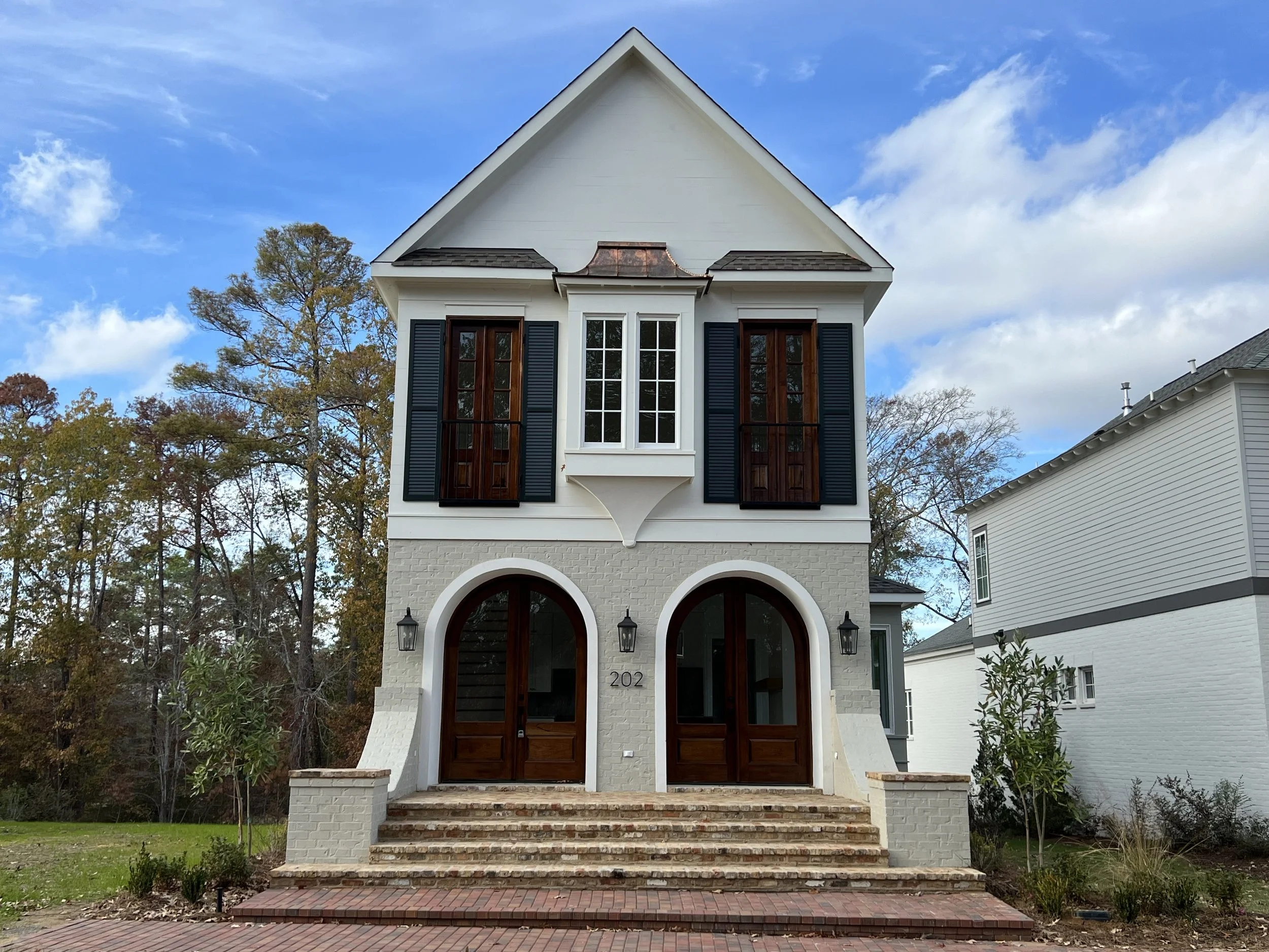 A modern three-story house with white exterior, dark wood accents, arched front doors, and black shutters, set against a partly cloudy sky and surrounded by trees and a brick-paved driveway.