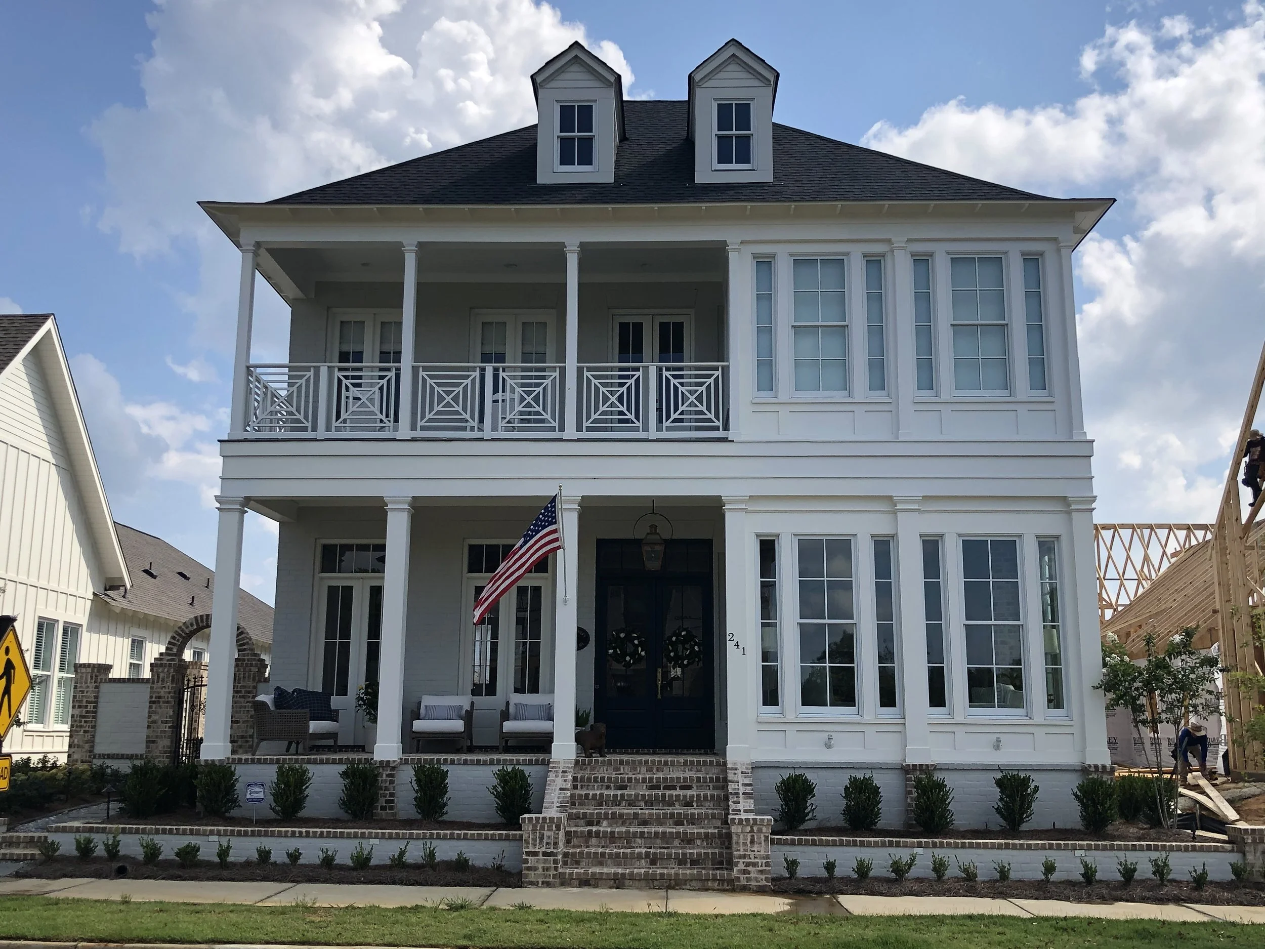 White multi-story house under construction with a front porch, American flag, and multiple large windows, with construction workers and a partly cloudy sky in the background.