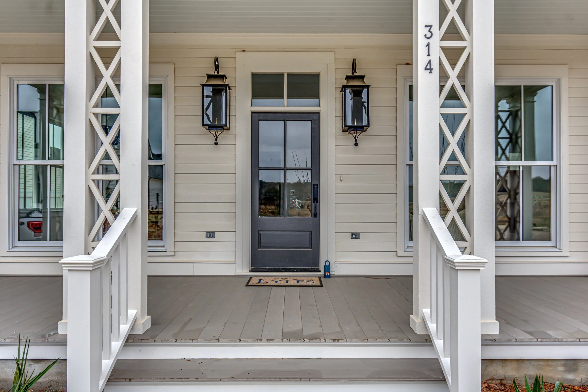 Front porch of a house with a black door, two lantern-style wall lights, and white lattice columns numbered 314. A welcome mat with the word "Lyles" is on the porch.