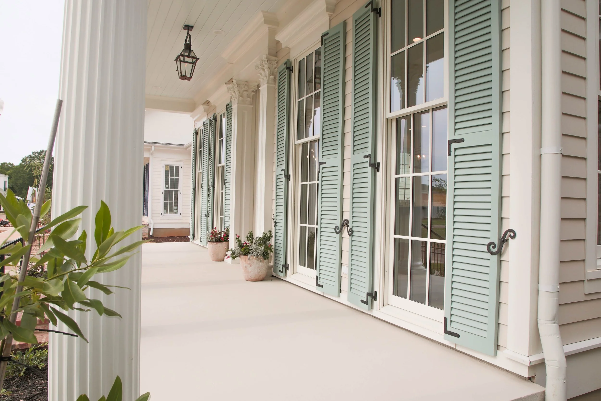 A porch area of a house with light blue shutters, white frames, potted plants, and decorative fixtures, with a white column on the left side.
