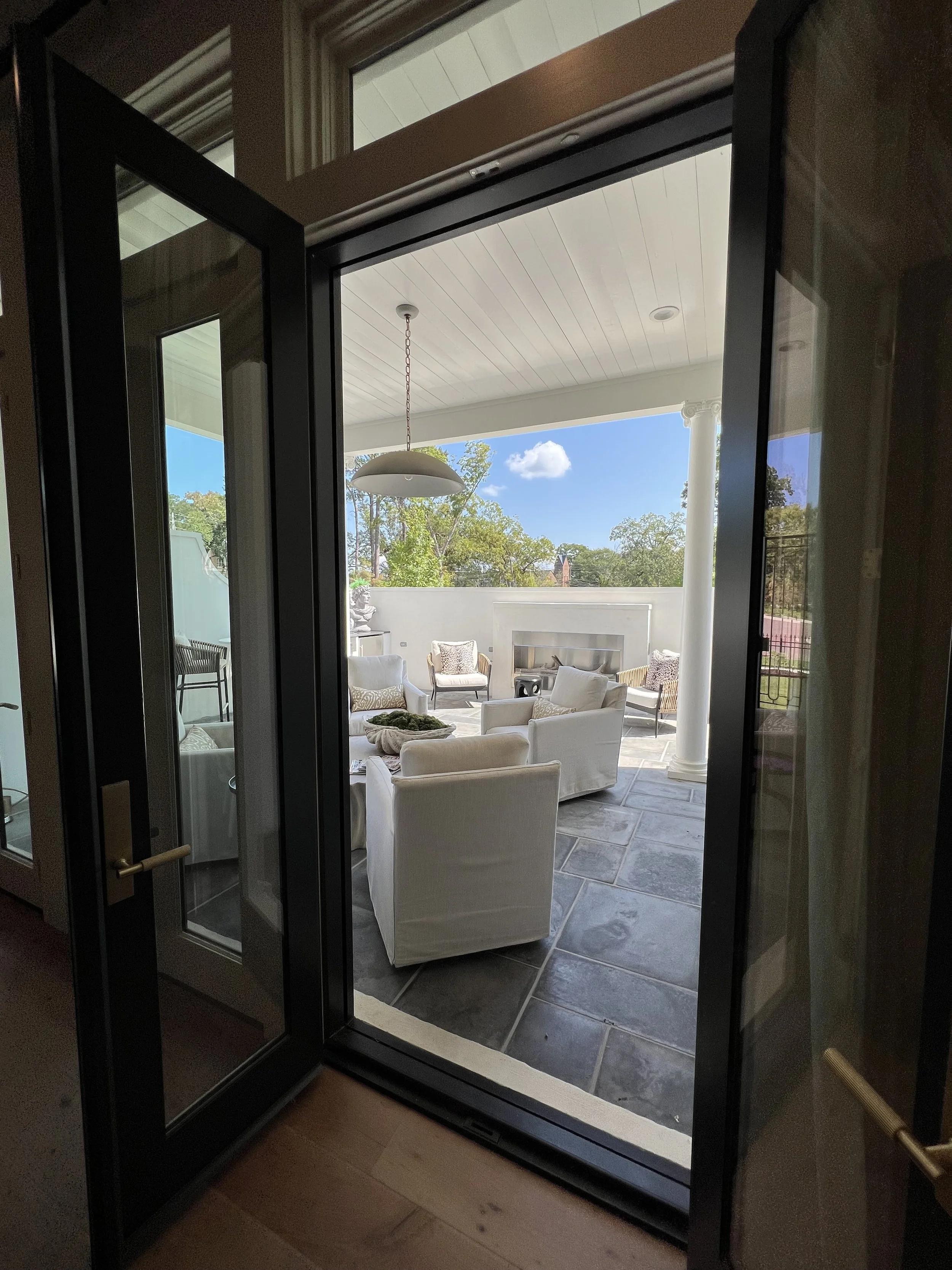 View through a glass door onto an outdoor patio with white armchairs, a fireplace, and trees in the background.