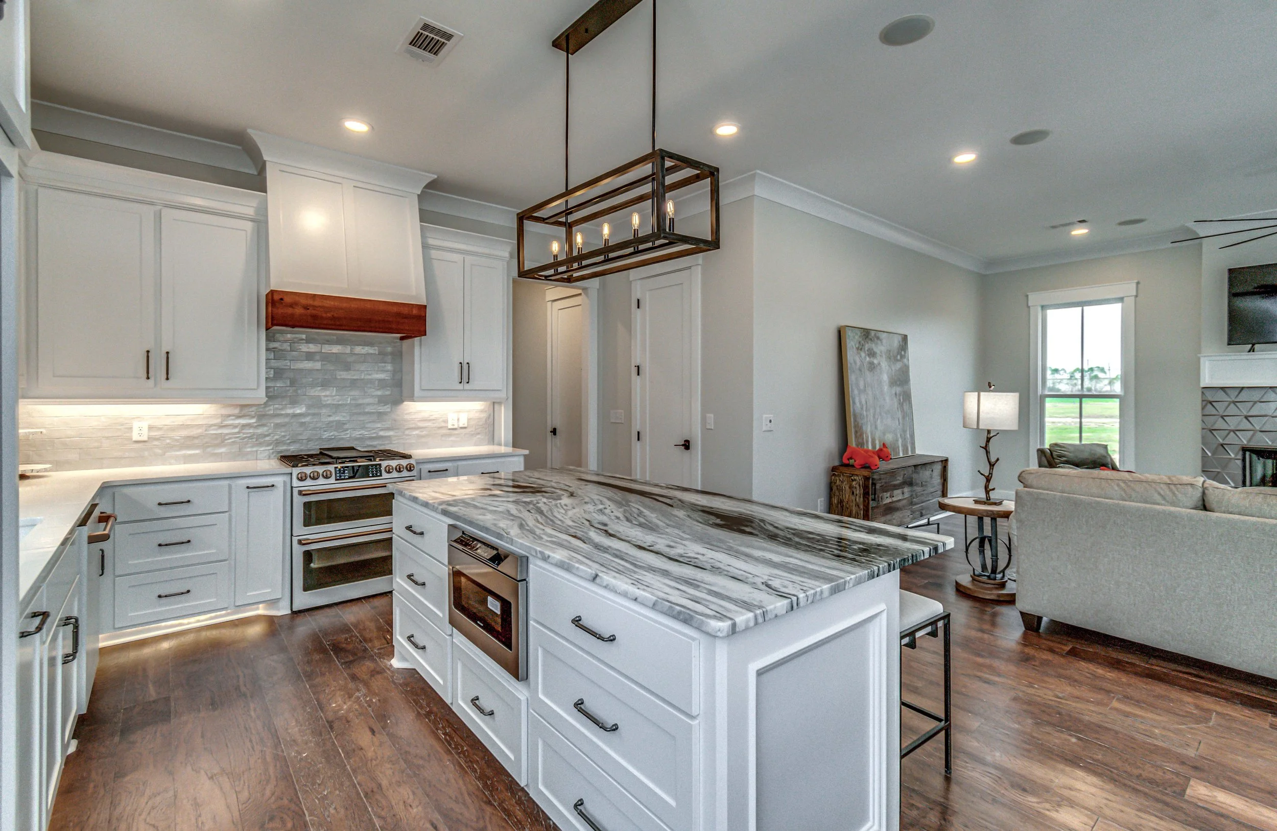 Modern open-concept kitchen with white cabinets, marble countertop island, stainless steel appliances, and wooden details, adjacent to a living room with beige sofas, a side table, and a window showing outdoor greenery.