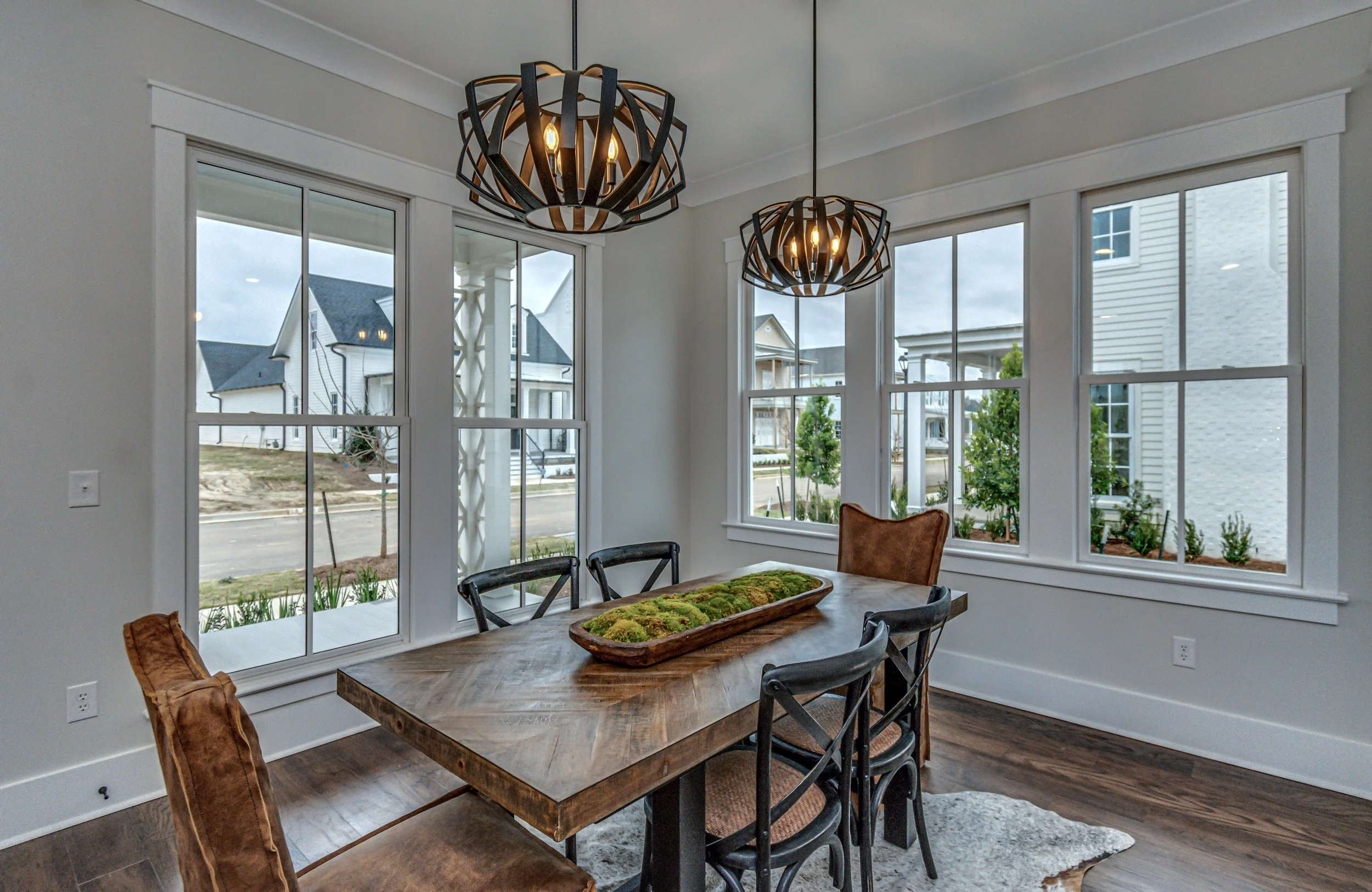 Dining room with wooden table, six chairs, two modern black and wood pendant lights, and large windows showing a neighborhood with houses and trees.