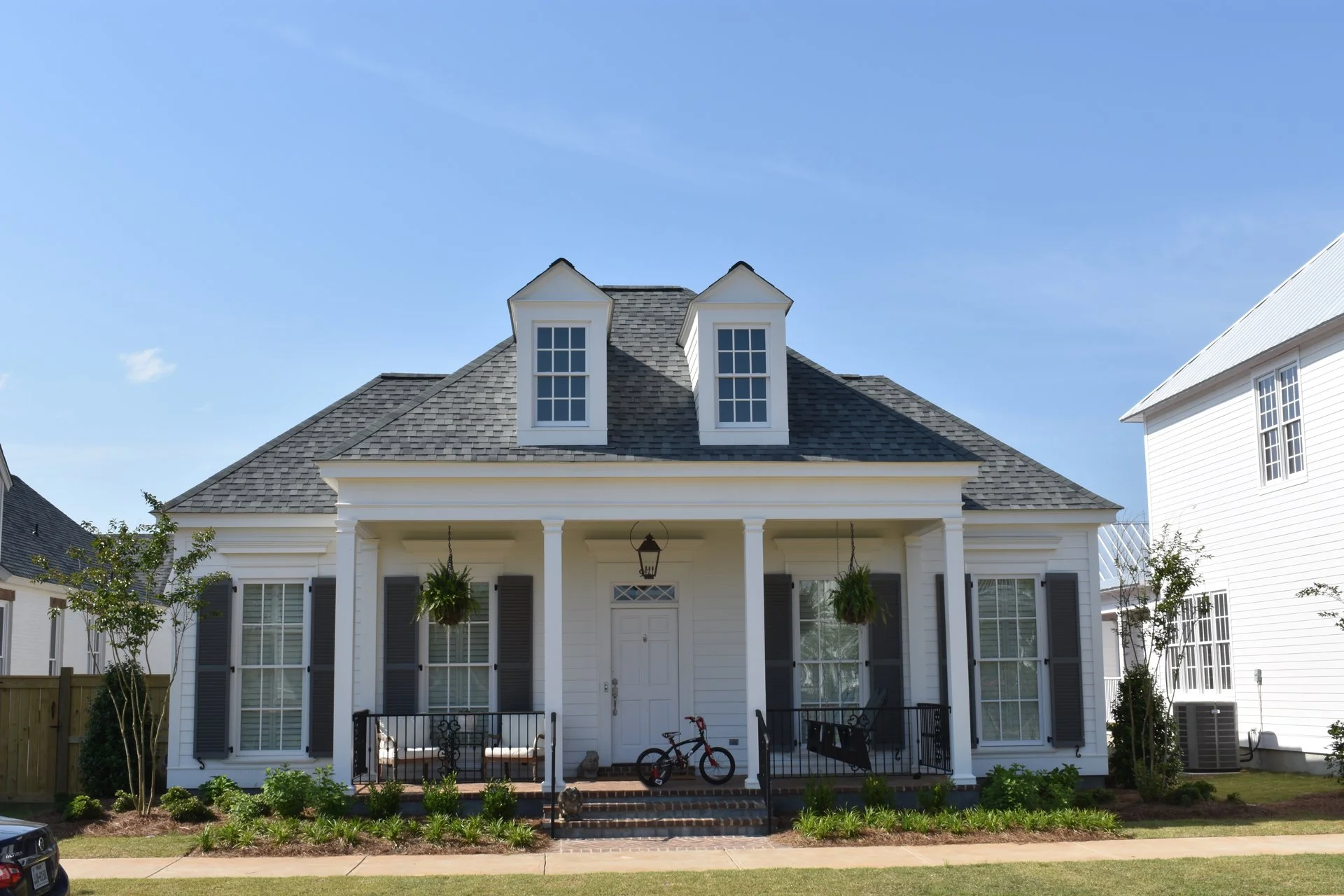 White house with gray roof, front porch with black railing, hanging plants, a bicycle, and a lawn with small bushes, under a clear blue sky.