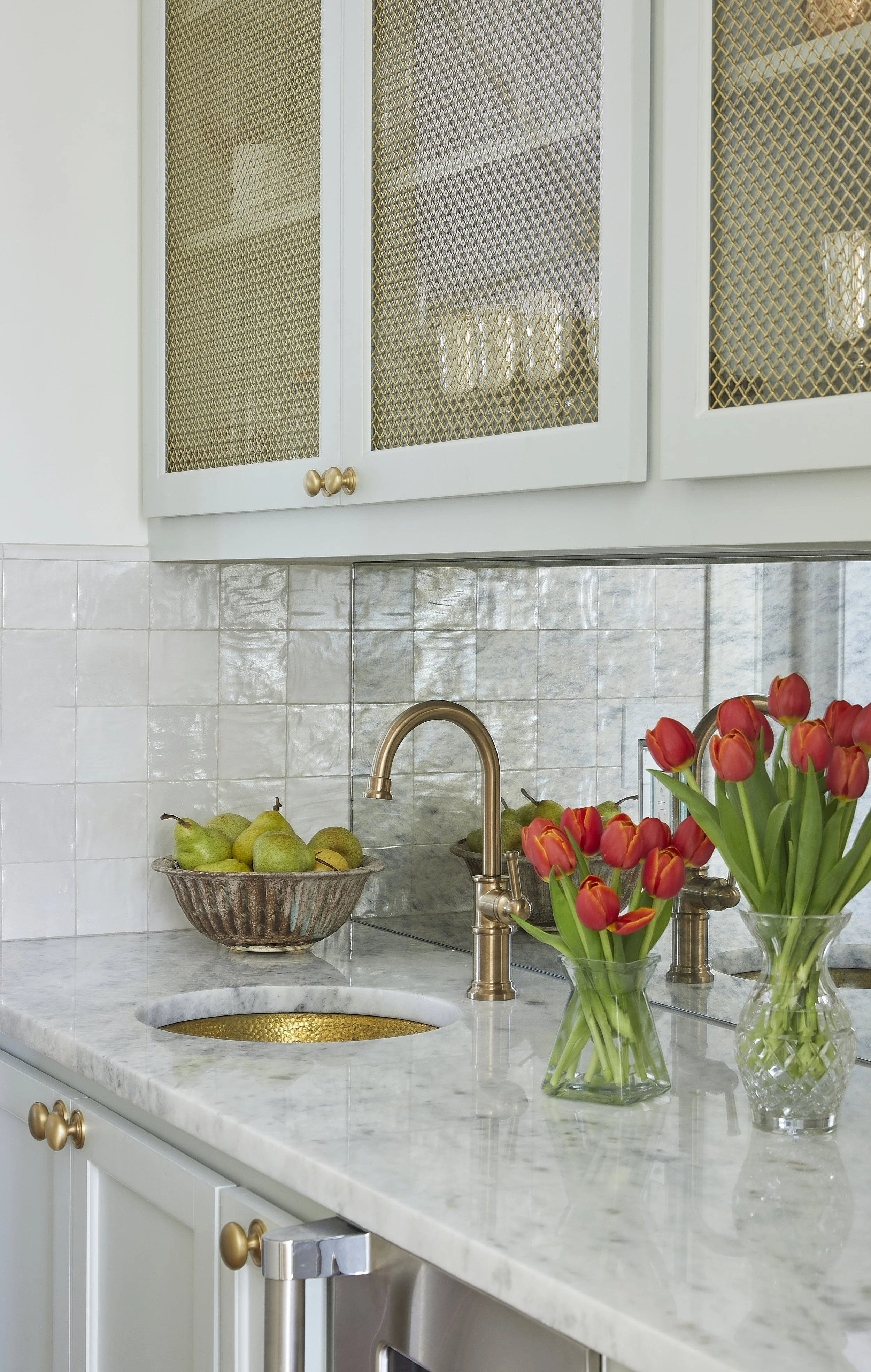 Kitchen countertop with a marble surface, a gold faucet, a vase with red tulips, a bowl of pears, and cream-colored cabinets with glass doors.