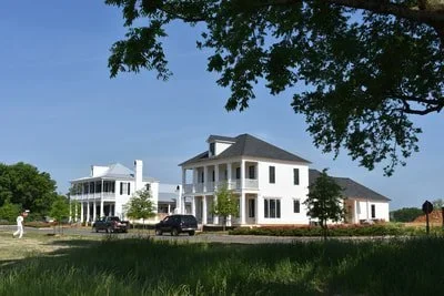 Two large white houses with front porches, situated in a green field with trees and cars parked nearby.