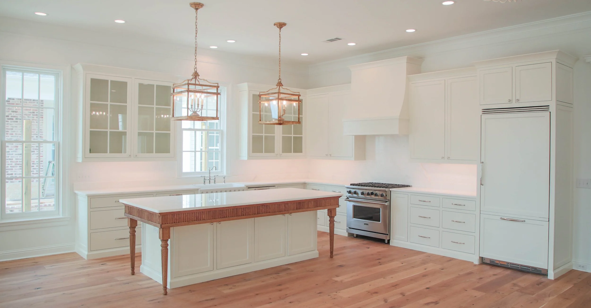Bright white kitchen with wooden accents, two pendant lights hanging over a central island, stainless steel stove, cabinetry, and hardwood floors.