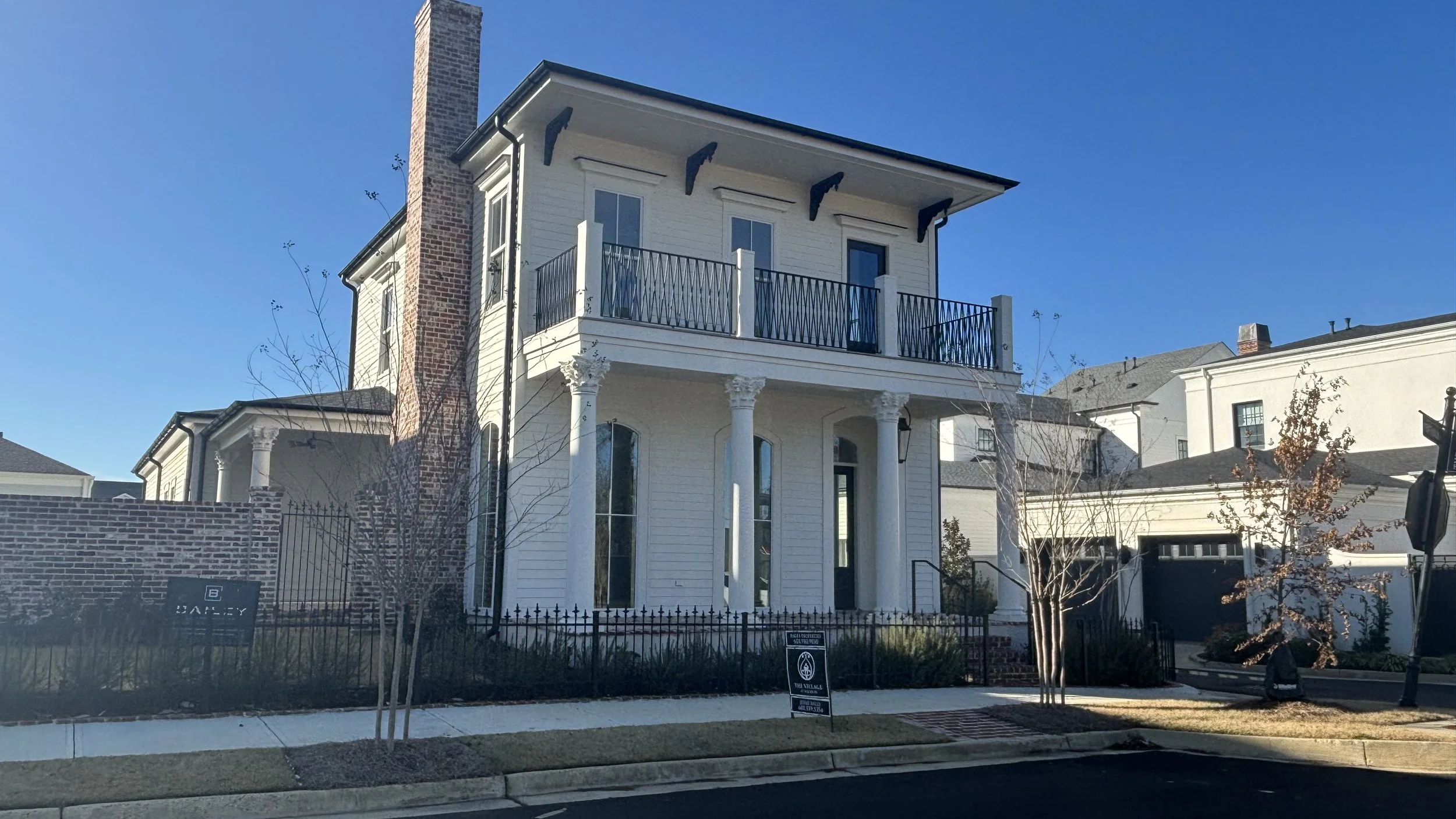 Two-story white house with tall columns in front, balcony on second floor, brick chimney on left side, black fence, and leafless trees