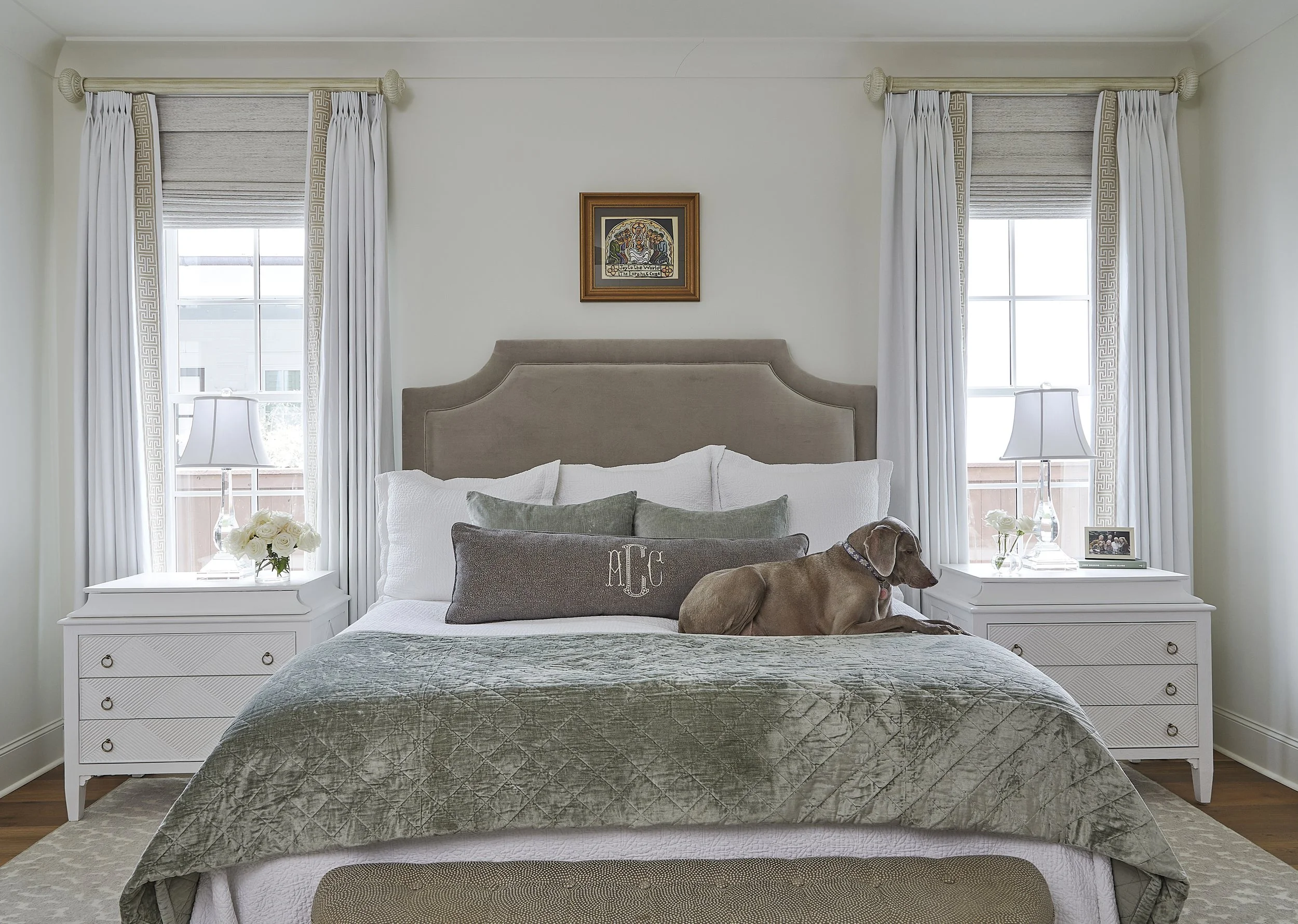 A bedroom with a beige upholstered headboard, white bedding, and decorative pillows, with a brown dog lying on the bed next to a window on each side with white curtains, and a framed picture on the wall above the headboard.