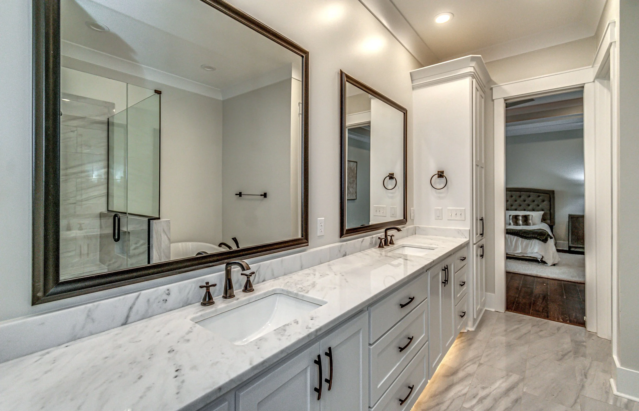 Bathroom with two large mirrors over a white marble countertop, double sinks, bronze faucets, in a spacious setting leading to a bedroom with a bed and wooden floor.