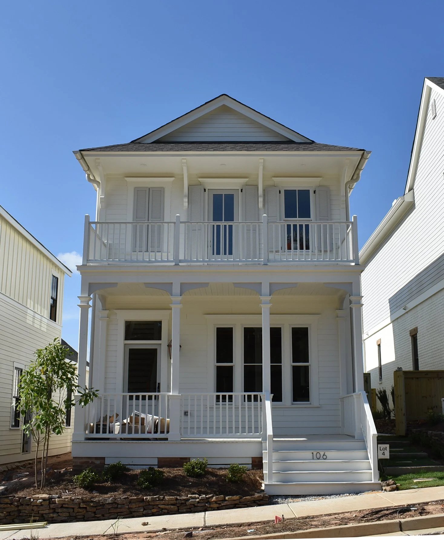 A three-story white house with a front porch and balcony, black roof, and nearby similar houses, under a clear blue sky.