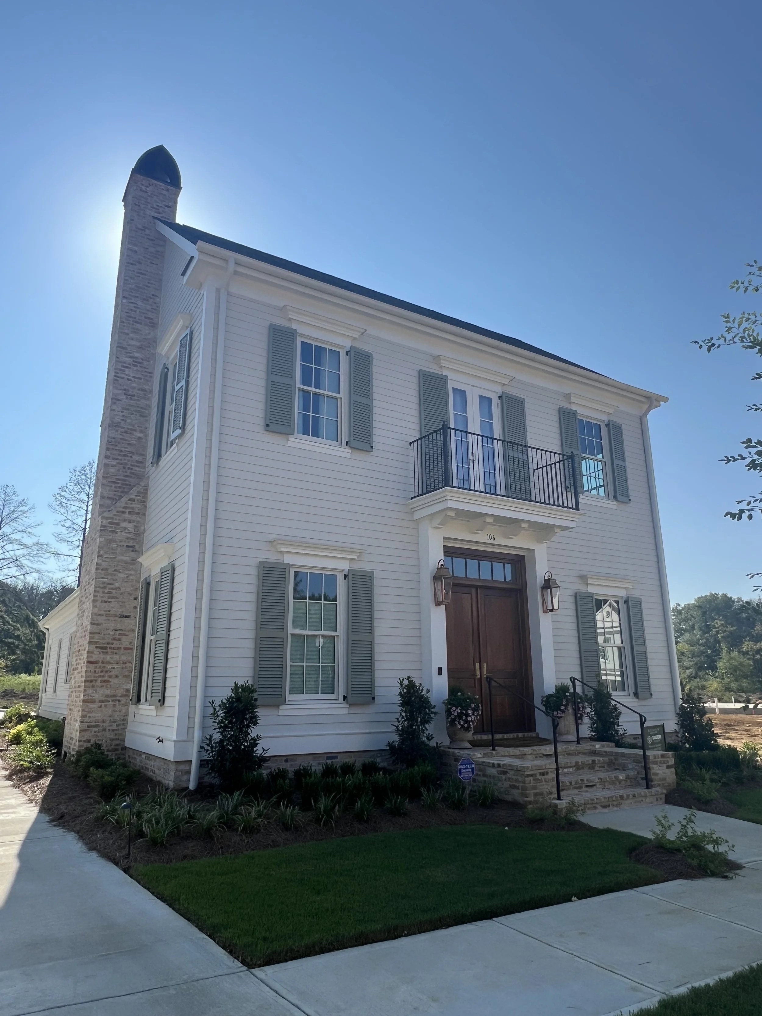 Front view of a two-story white house with gray shutters, a small balcony, stone steps leading to the front door, well-maintained garden, and a chimney on the left side. The sky is clear and blue.