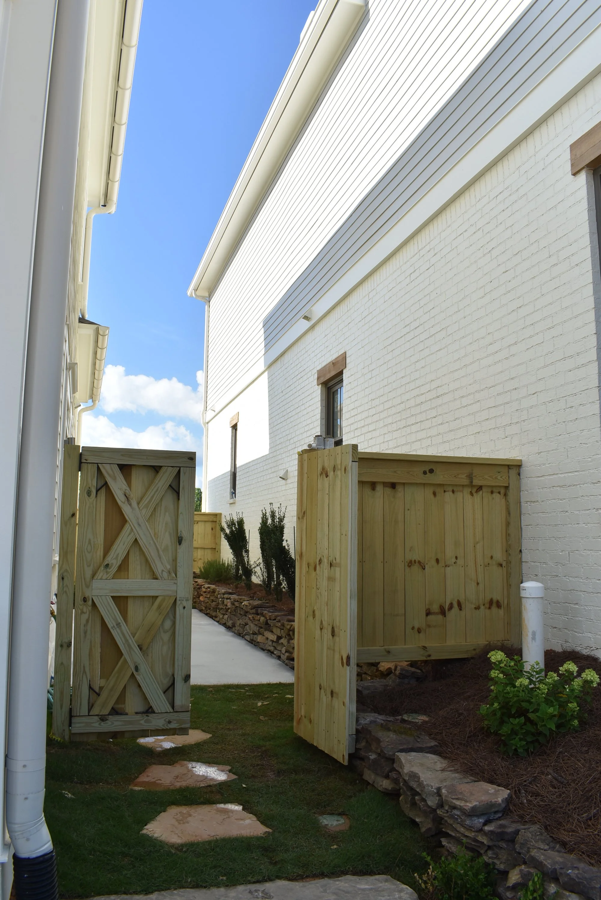 View of a residential backyard with a white brick house, a small garden with bushes and rocks, a stone pathway, and a set of wooden gates.