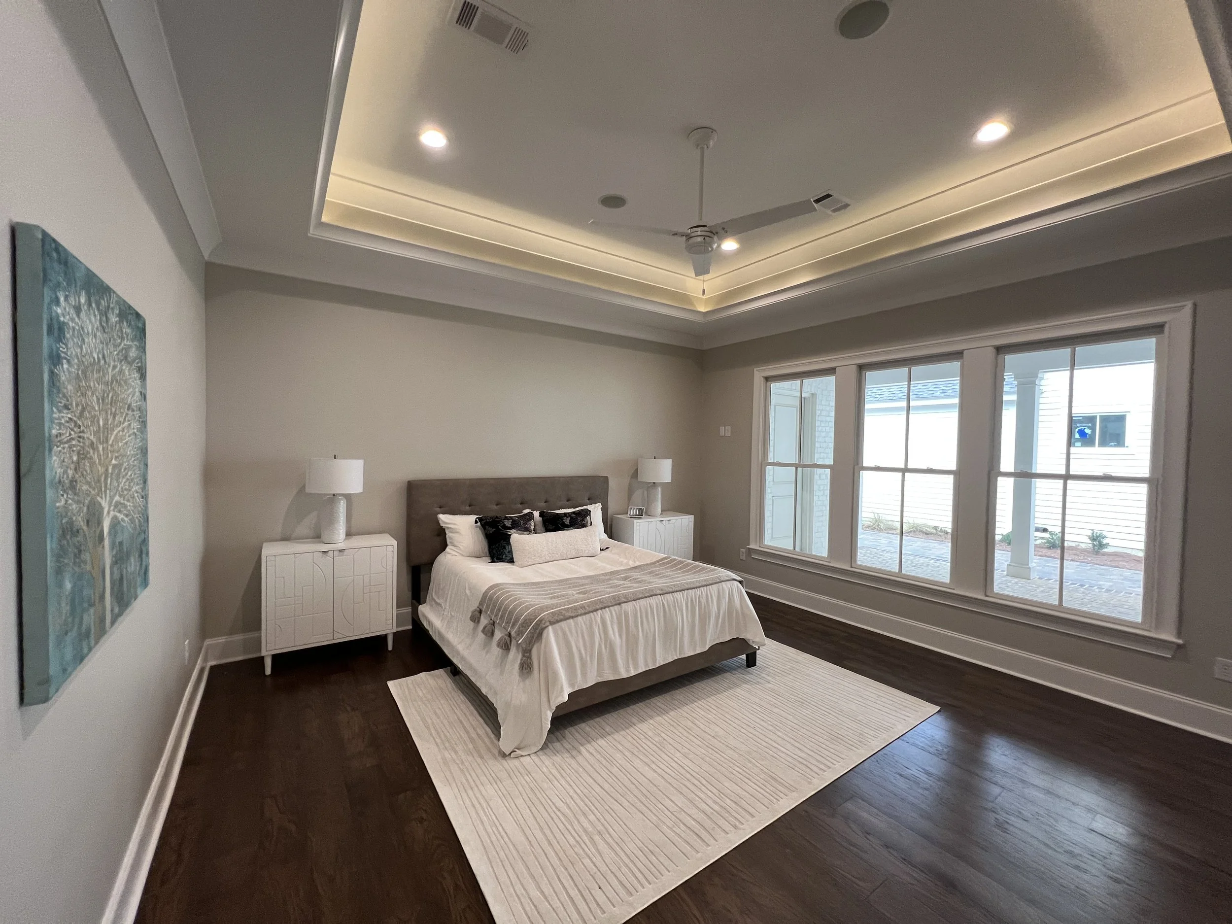 Modern bedroom with a bed, nightstands, lamps, painting, large windows, hardwood floor, and a ceiling with recessed lighting and a ceiling fan.