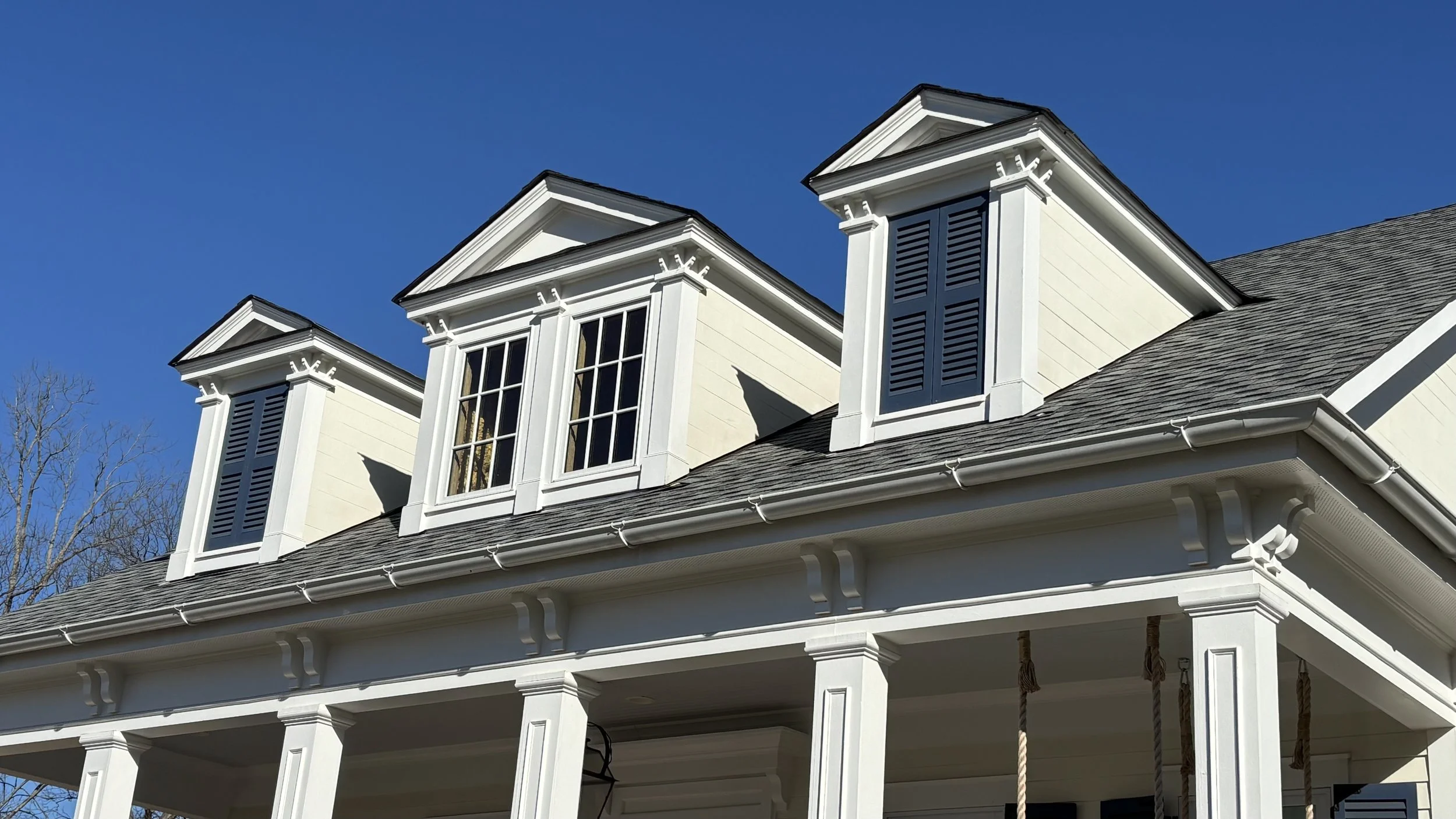 Close-up of a white house with multiple dormer windows, gray shingle roof, and decorative trim, against a clear blue sky.