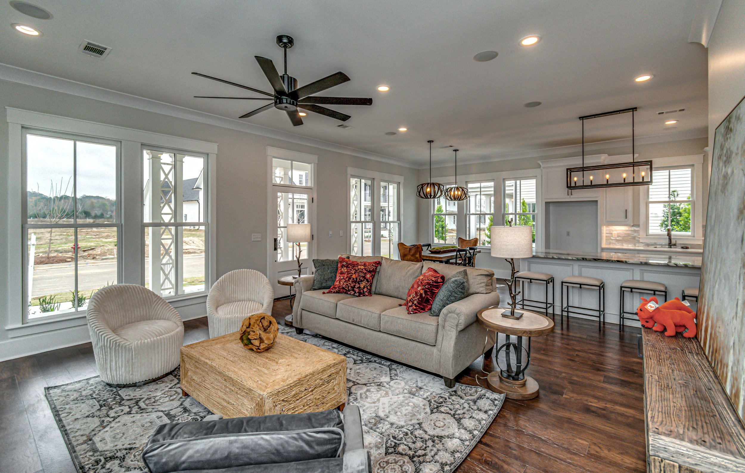 Open-concept living room with large windows, a ceiling fan, a gray sofa with decorative pillows, two cream-colored accent chairs, a woven coffee table, a patterned area rug, a side table with a sculpture, and a view of a bright, modern kitchen with b