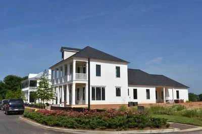 A large white residential house with a dark roof, multiple balconies, and surrounding greenery, under a clear blue sky.