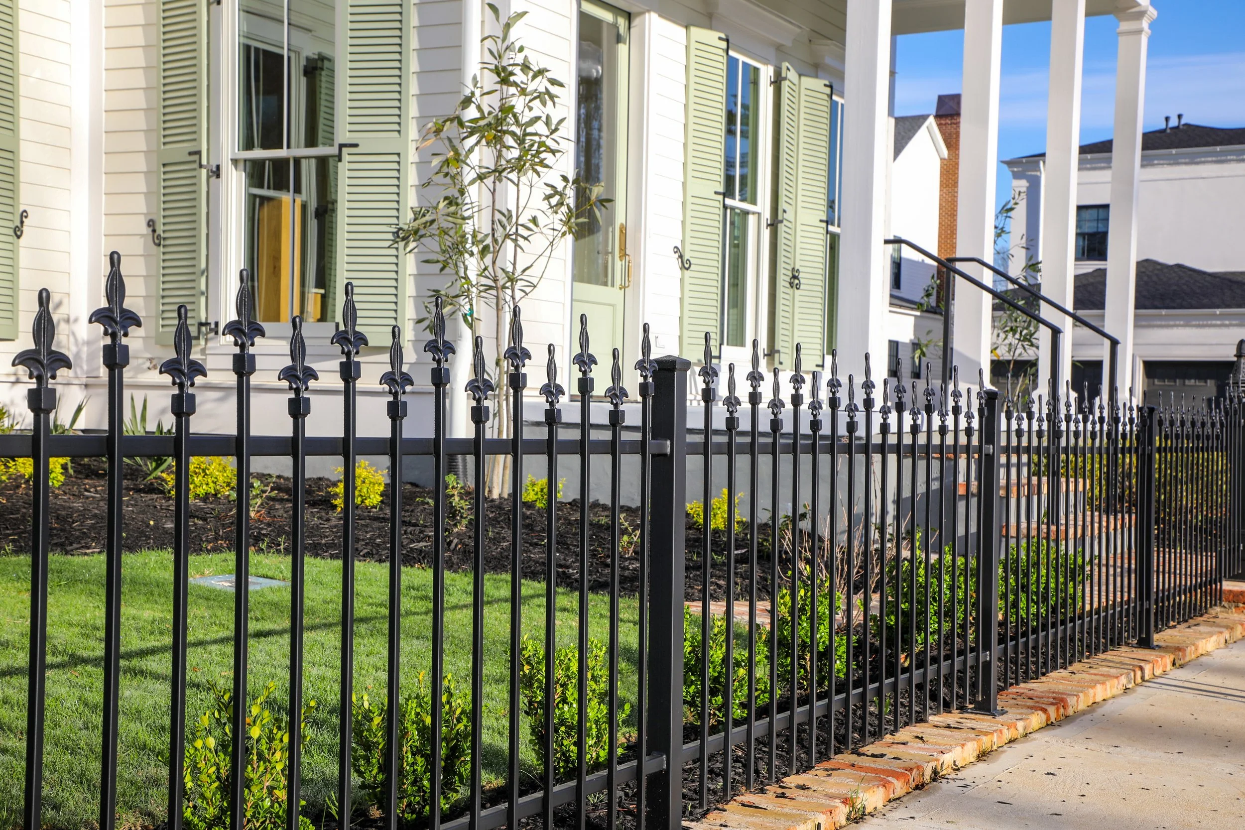 A house with white walls, green window shutters, and a front porch surrounded by a black metal fence with brick foundation. There are small bushes and plants in the garden area in front of the house.
