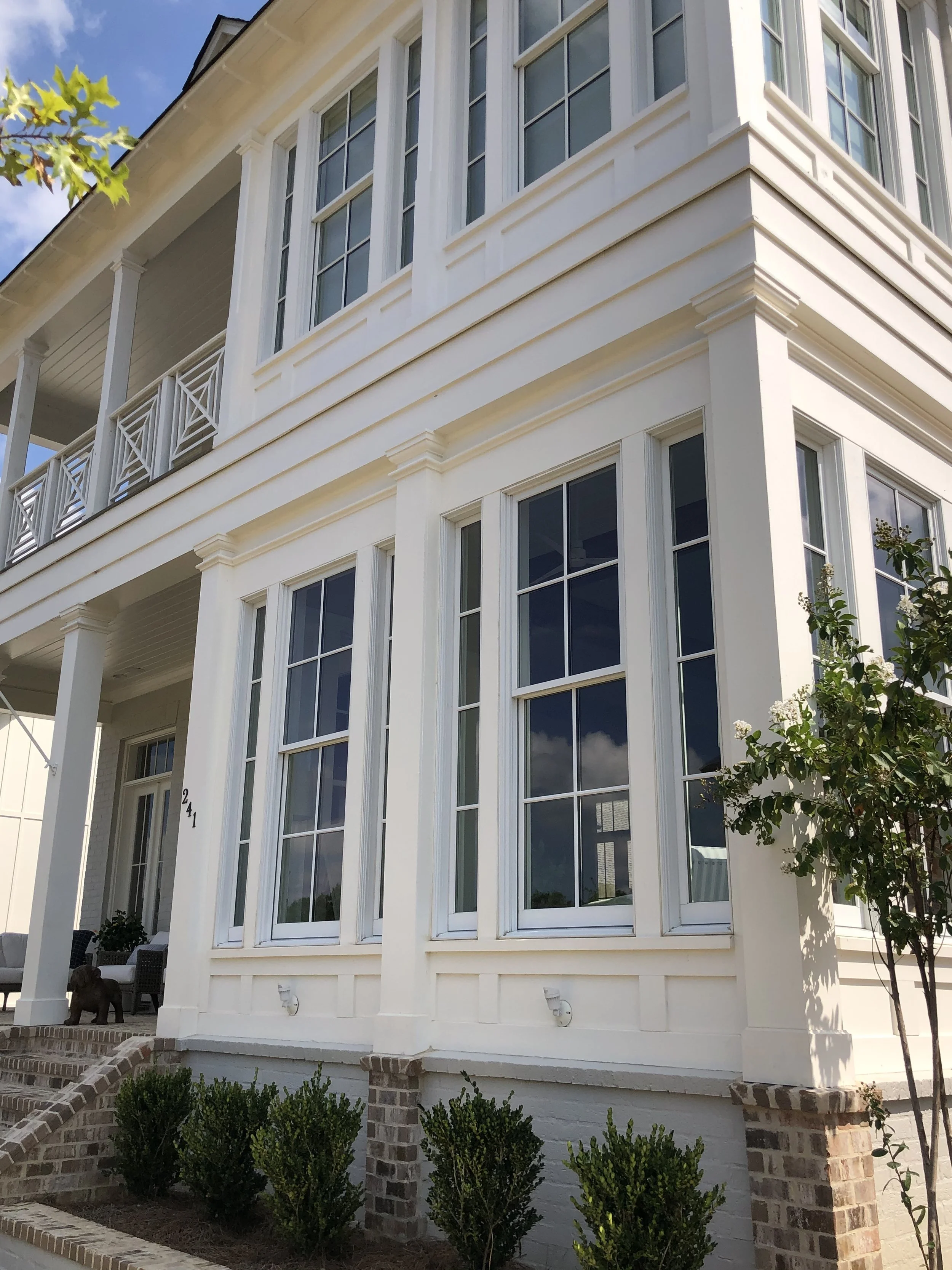 Close-up of a large white house with multiple large windows, brick pillars supporting a porch, and landscaped greenery in front.