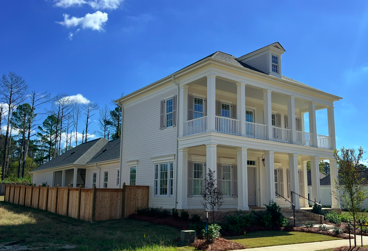 A large, white, three-story house with a wraparound porch, multiple windows, and a pitched roof. The house is surrounded by a wooden fence and has small trees and landscaping in the front yard, with a bright blue sky and some clouds overhead.