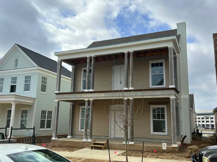 Side view of a two-story house under construction with exposed wooden columns and unfinished exterior, neighboring houses visible, and a small leafless tree in front.