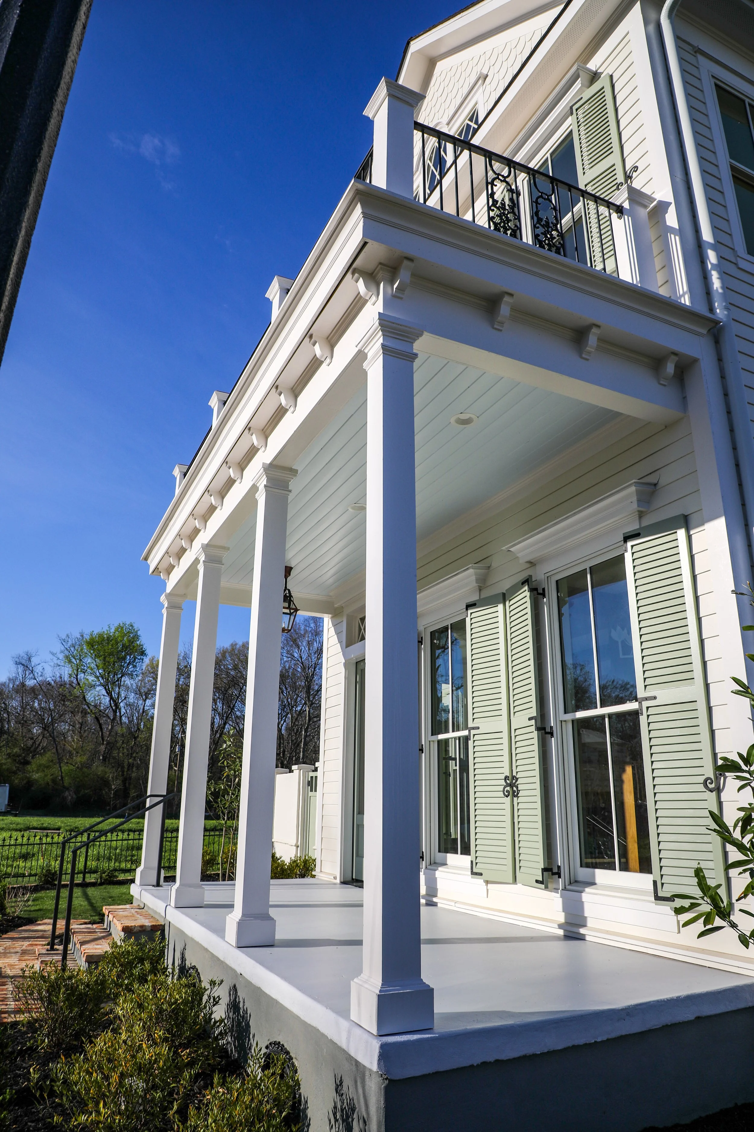 White porch with columns, railings, and green shutters on a house, under a clear blue sky.