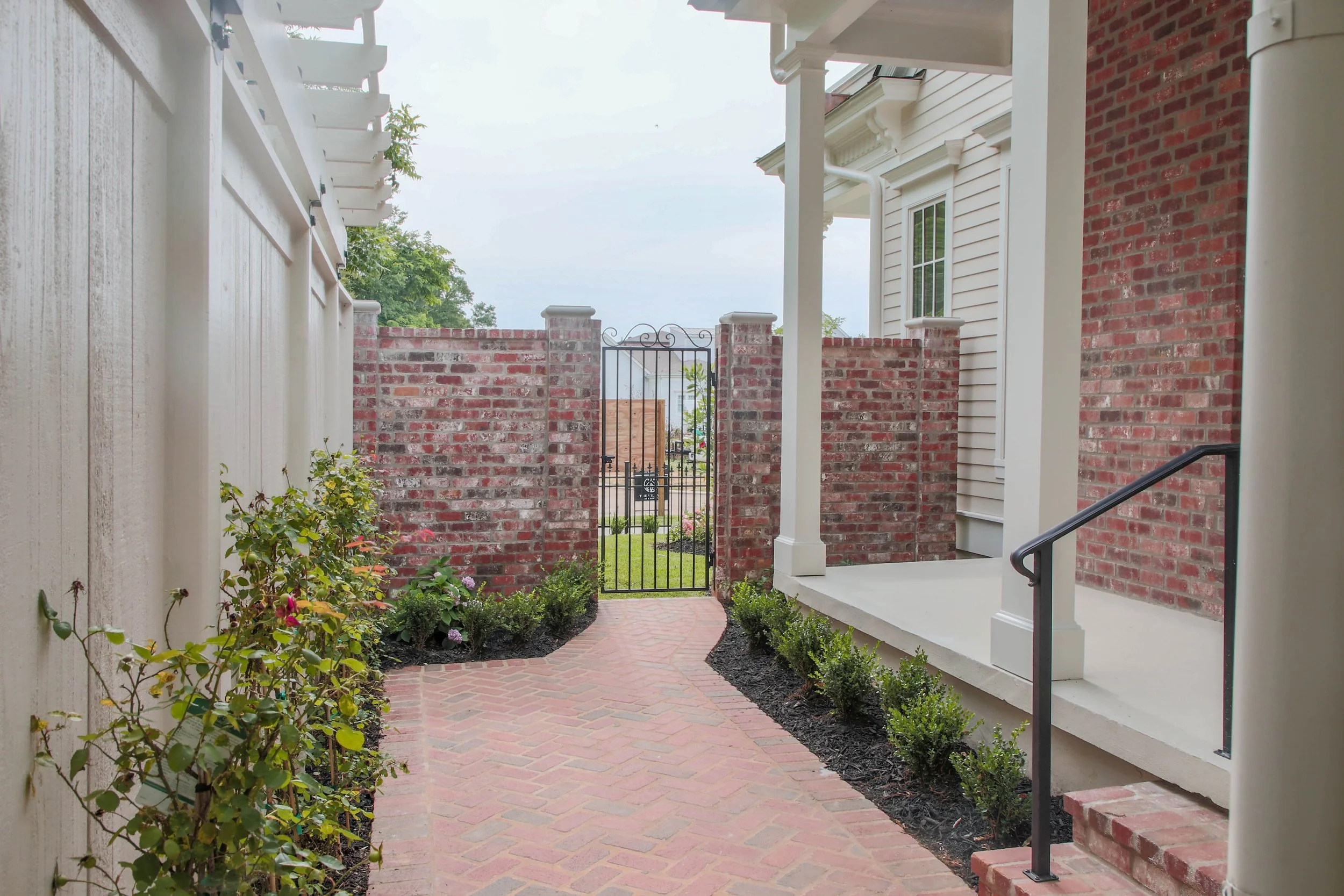 A narrow brick pathway lined with small bushes on the right and a garden with flowers on the left leading to a black wrought iron gate. The scene is enclosed by brick and siding walls of a house, with a porch on the right side.