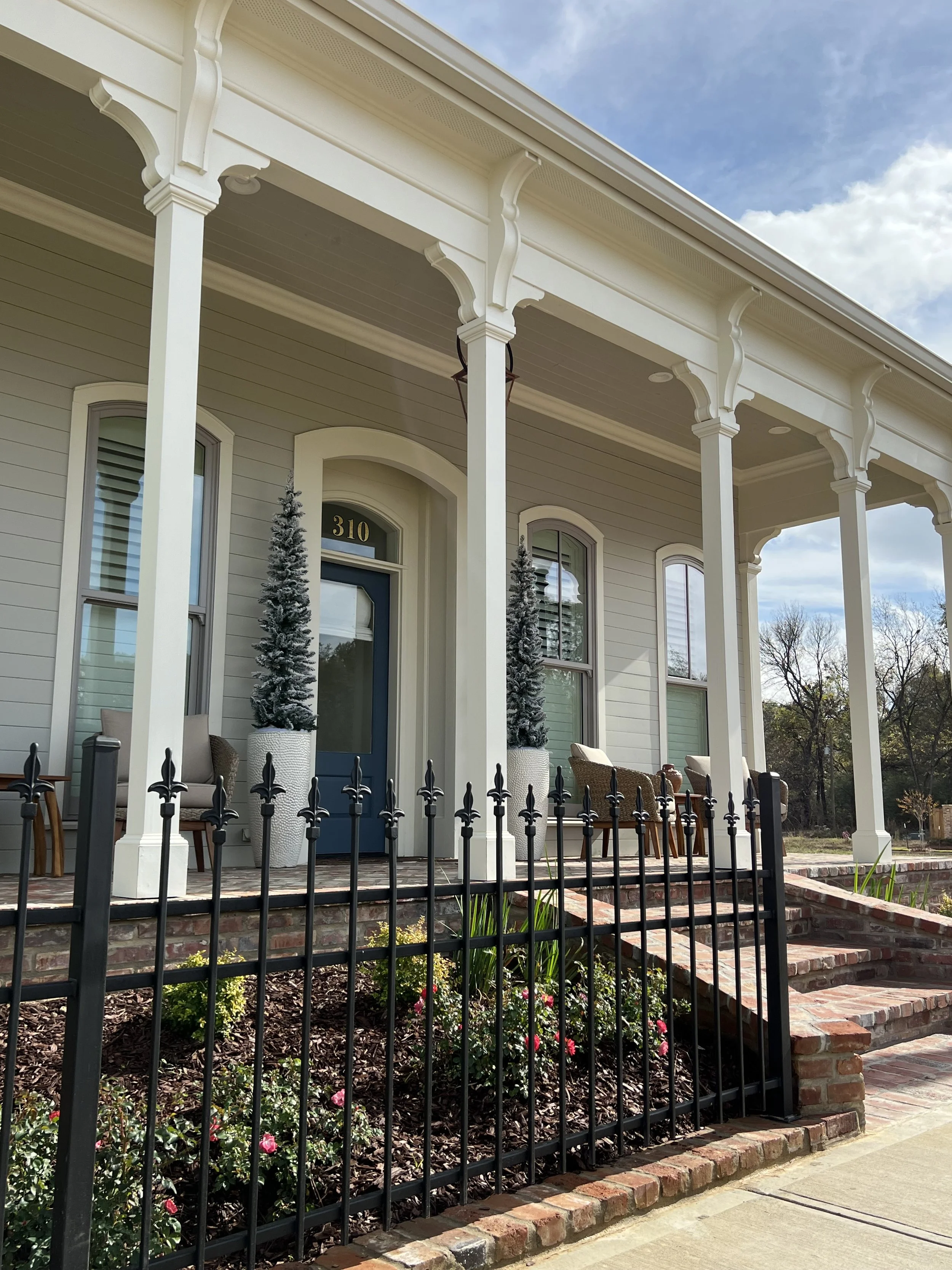 Front porch of a house with beige siding, white trim, decorative columns, and a blue door with the number 310. There are two potted mini Christmas trees, outdoor furniture, and a black wrought iron fence with brick steps leading up to the porch. The 