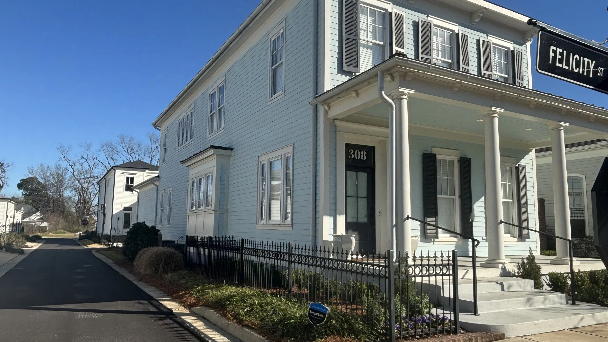 A white, two-story house with black shutters located on a corner with a sign that reads Felicity Street, number 308, featuring a front porch with columns, a small ramp, and a black metal fence along the sidewalk.
