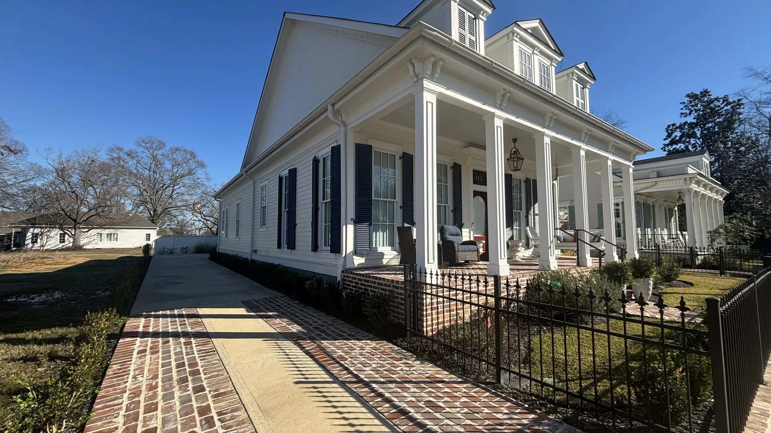 Front view of a white house with black shutters, brick stairs, porch furniture, and a black fence, under a clear blue sky.