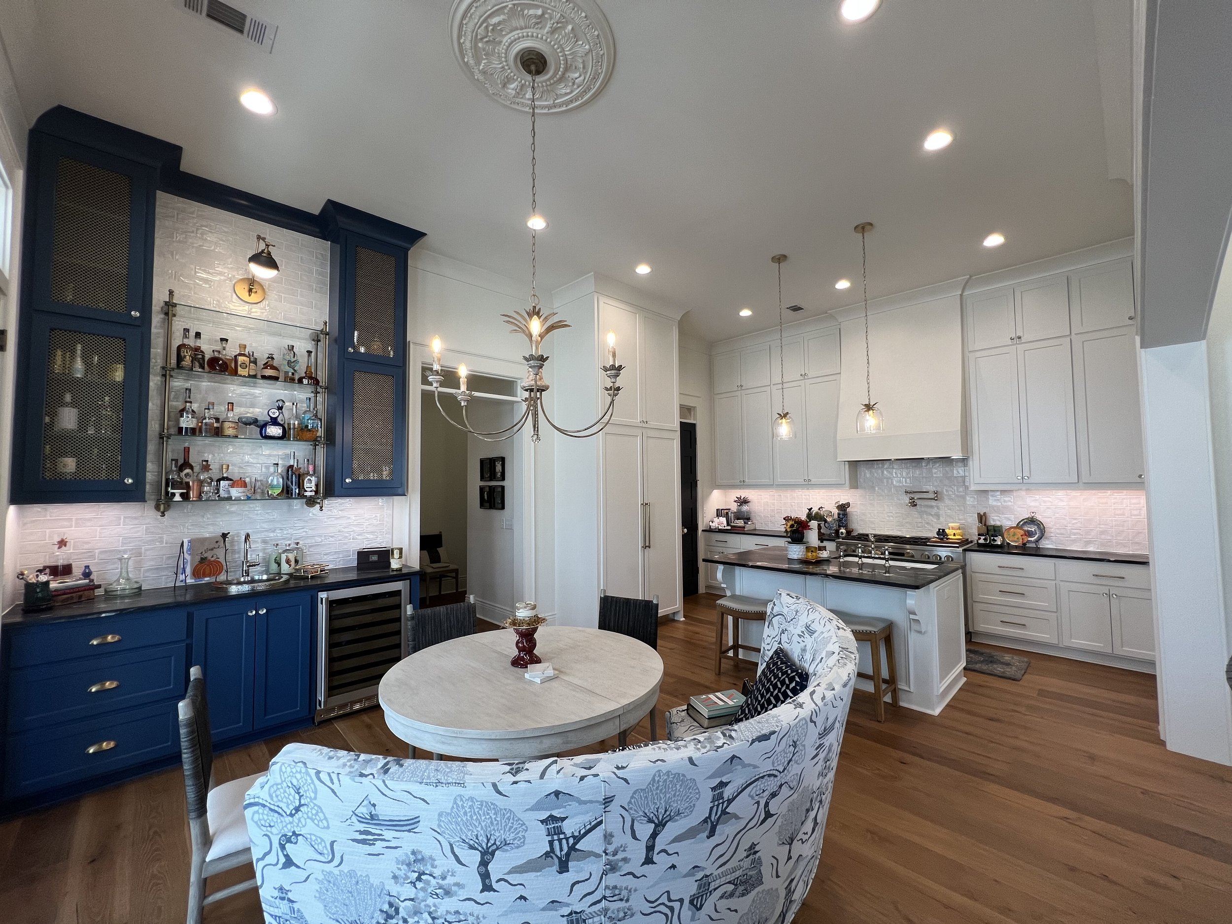 Bright kitchen with white cabinets, a central island, and a wall-mounted blue bar cabinet with glass shelves displaying liquor bottles. There is a chandelier, pendant lights, and a round dining table with assorted chairs.