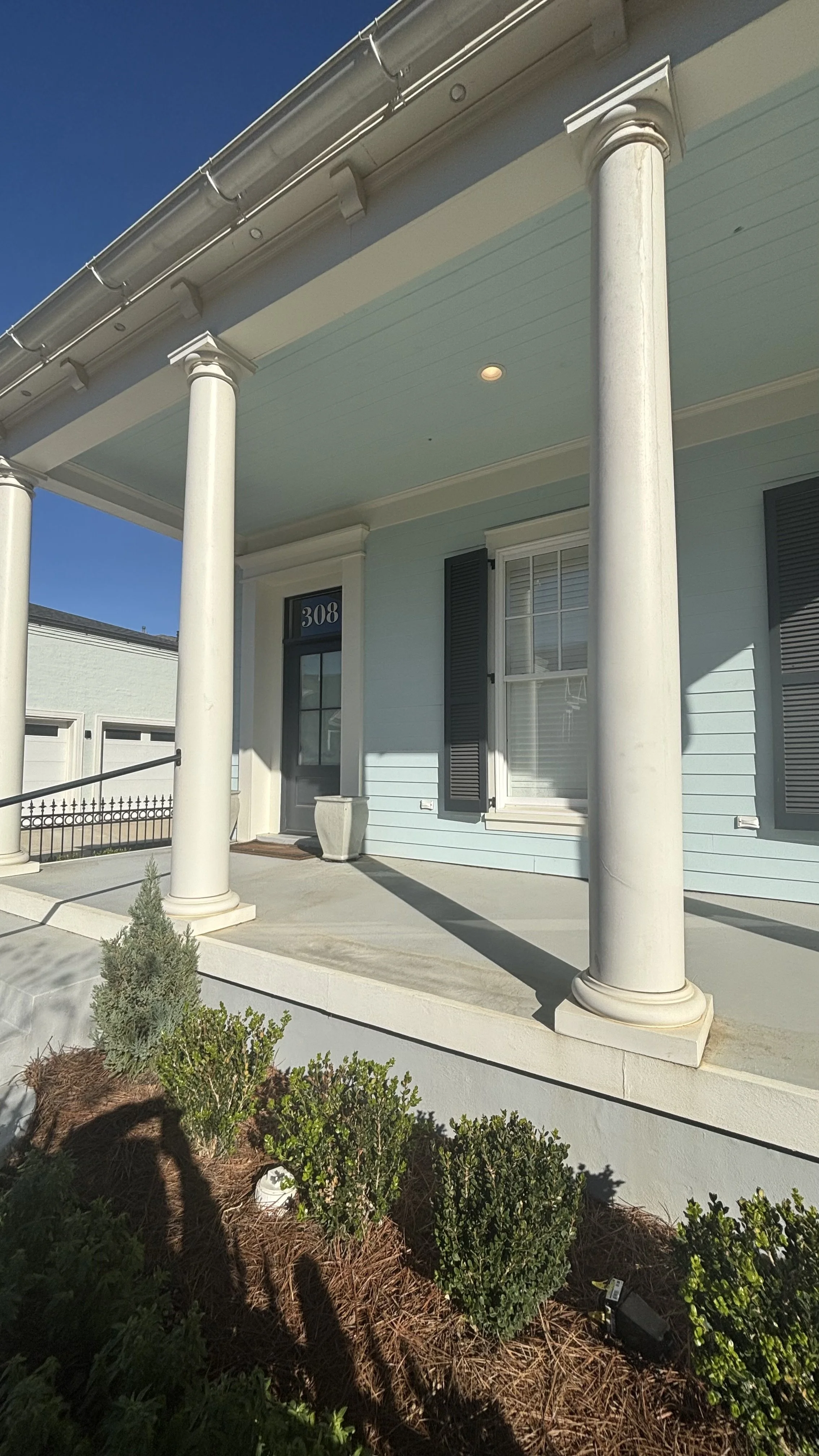 Front porch of a house with light blue exterior, white columns, black shutters, and a door with the number 308, sunlight casting shadows, small plants in front.