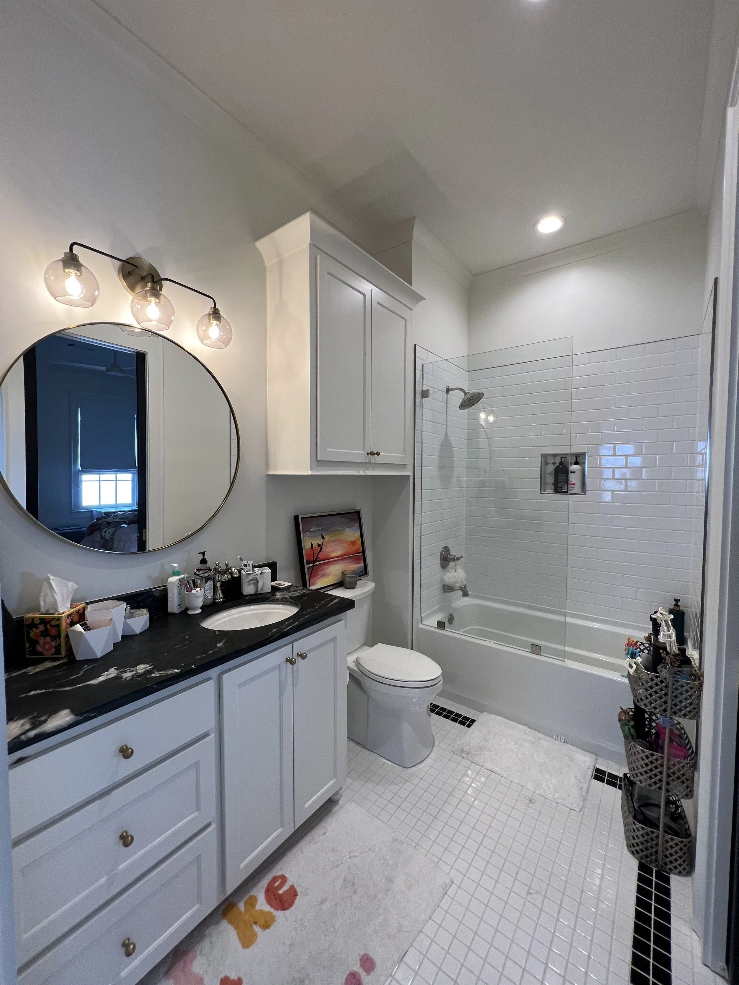 Modern white bathroom with a black countertop vanity, round mirror, wall-mounted light fixture, toilet, bathtub with glass shower screen, and tiled walls and floors. There are towels and toiletries visible.