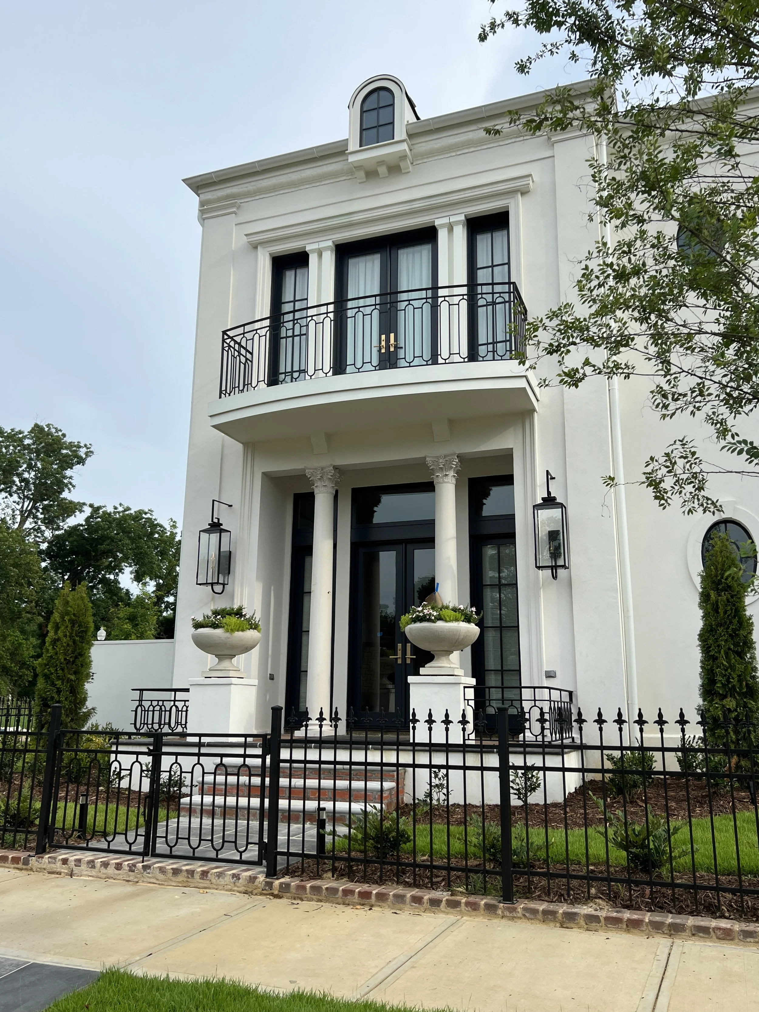 Front view of a white, three-story house with black doors and black window frames, decorative columns at the entrance, a small balcony with black metal railing, large outdoor lanterns, and two white planters with flowers on either side of the entranc